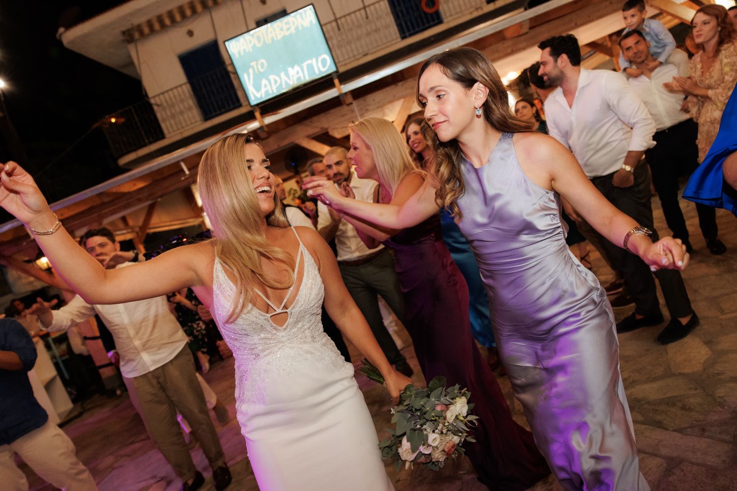 Bride and bridesmaid dancing joyfully together at an outdoor evening wedding reception.