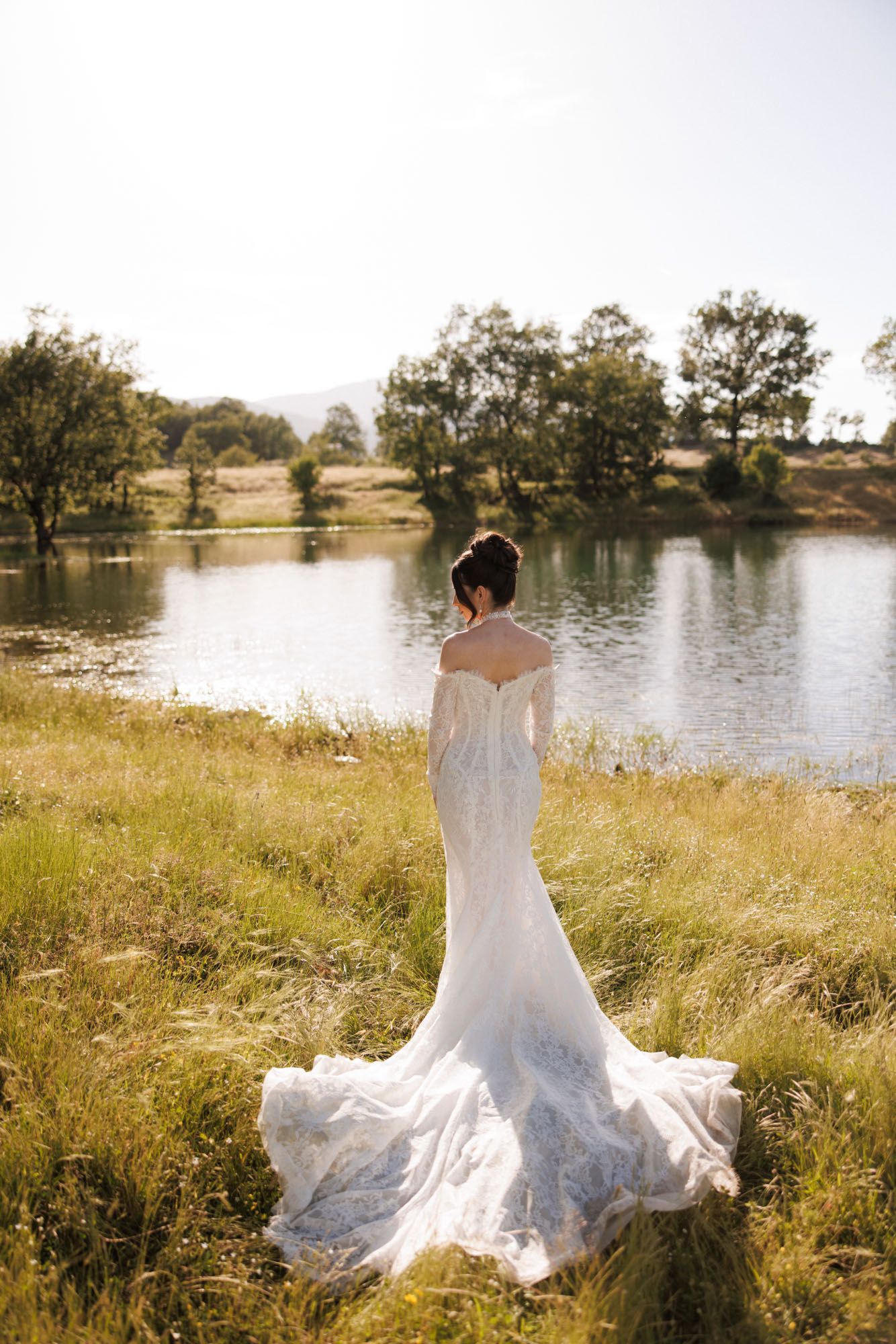 Bride in off-shoulder lace gown with train stands by a serene lake surrounded by lush greenery on a sunny day.