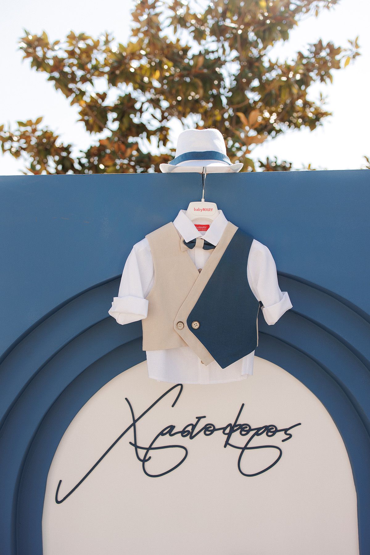A small boy's christening outfit with white shirt, beige vest, and hat displayed on a blue decorative backdrop.