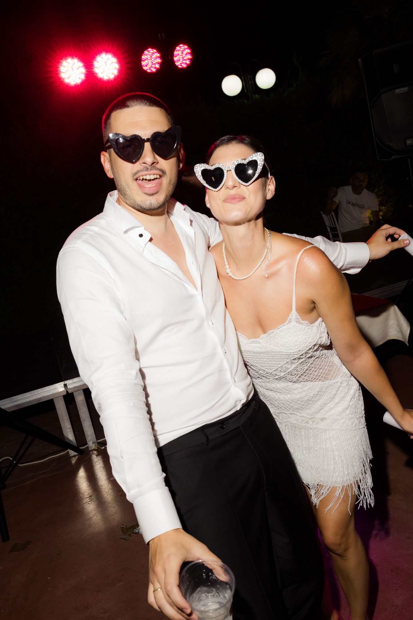 Two people in white outfits and heart-shaped sunglasses posing together at a nightclub party with colorful stage lights.
