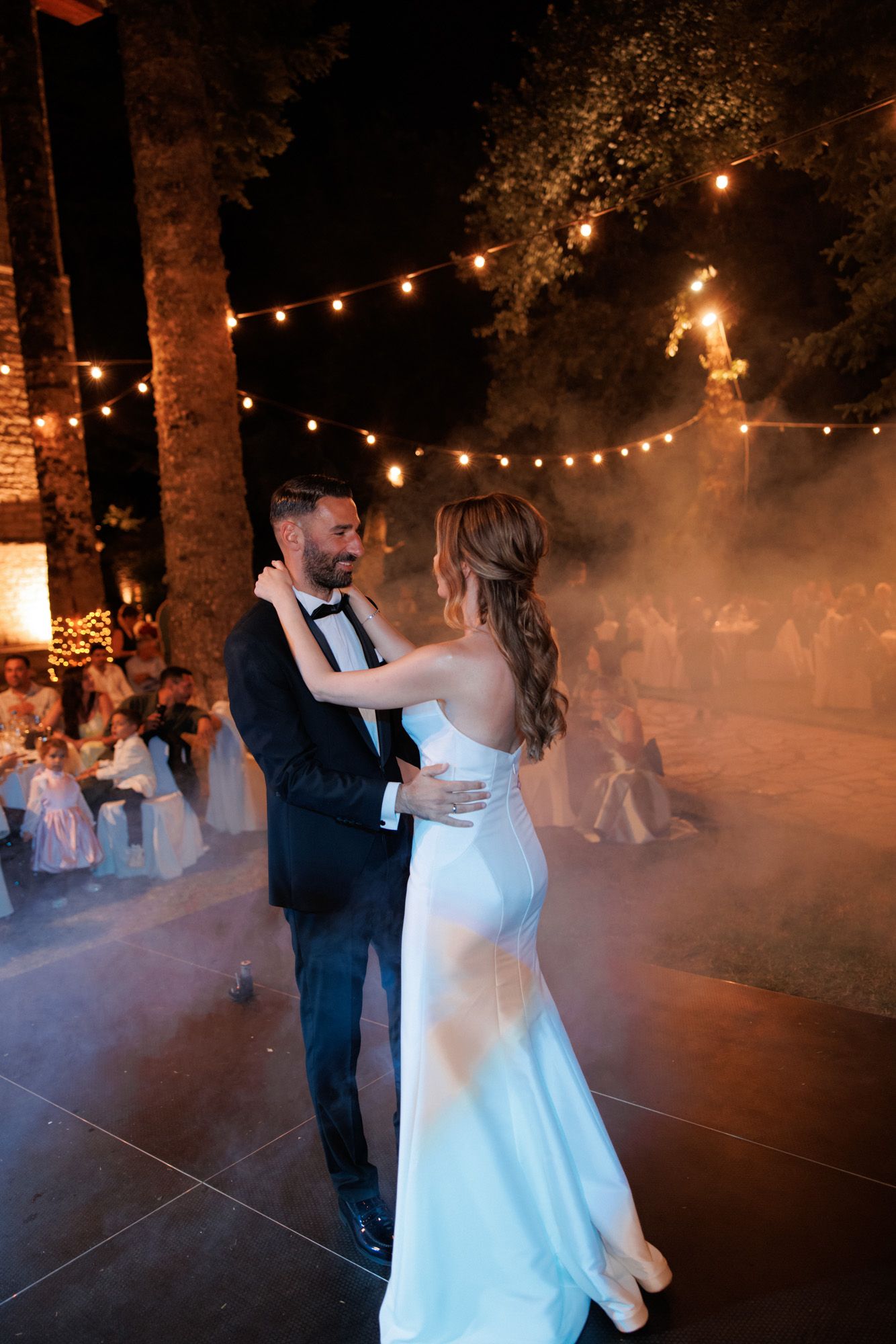 Bride and groom share first dance on outdoor dance floor surrounded by string lights and atmospheric fog.