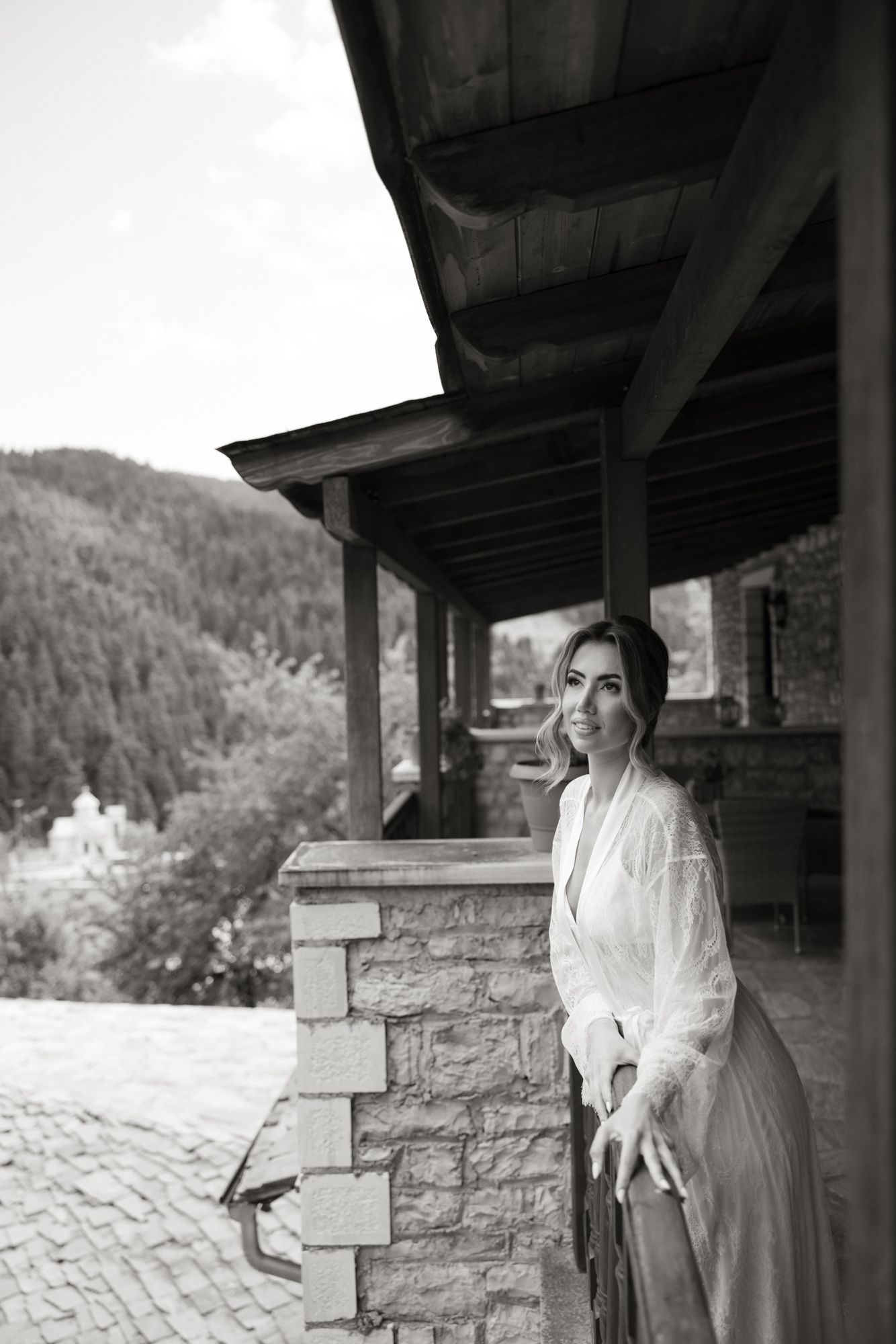 Black and white portrait of smiling bride in white robe standing on stone balcony with mountain view.