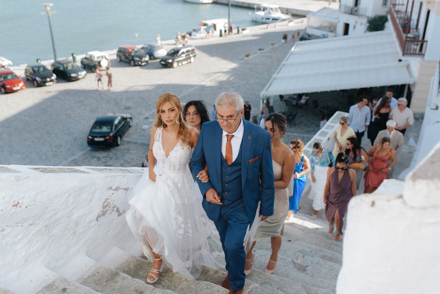 Bride in white gown walks up stone steps with groom in blue suit at a seaside wedding venue.