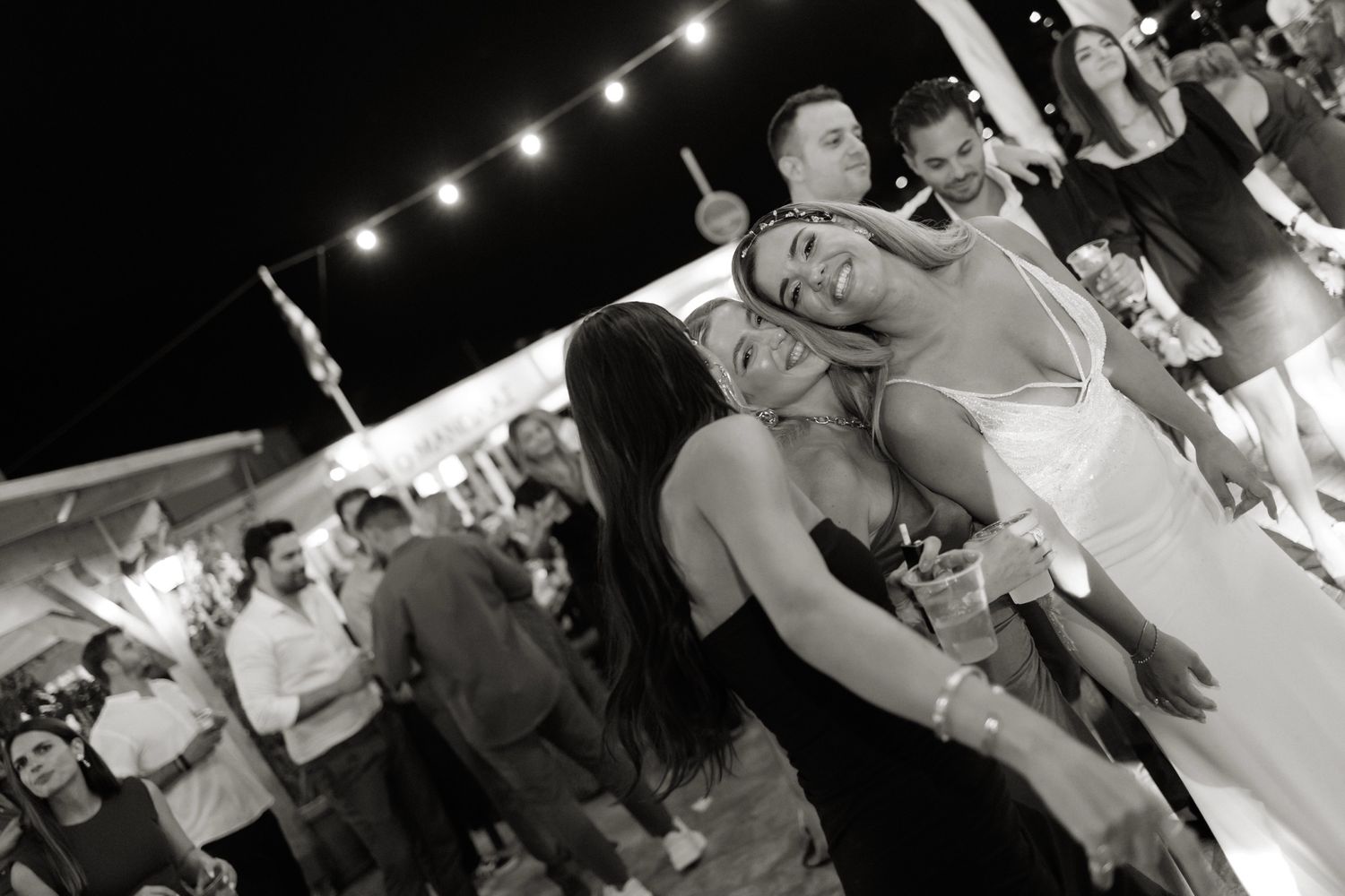 Bride embraces bridesmaid warmly on crowded outdoor wedding dance floor in black and white photo.
