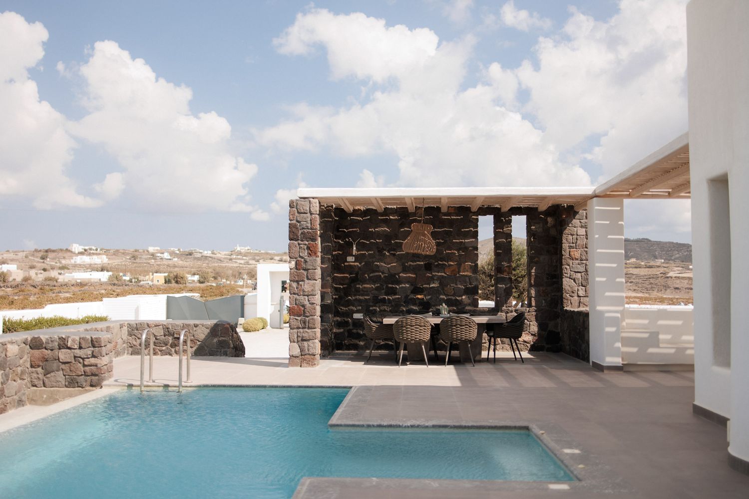 Luxury villa with a sparkling pool and stone pergola dining area under a cloudy sky in Mykonos, Greece.