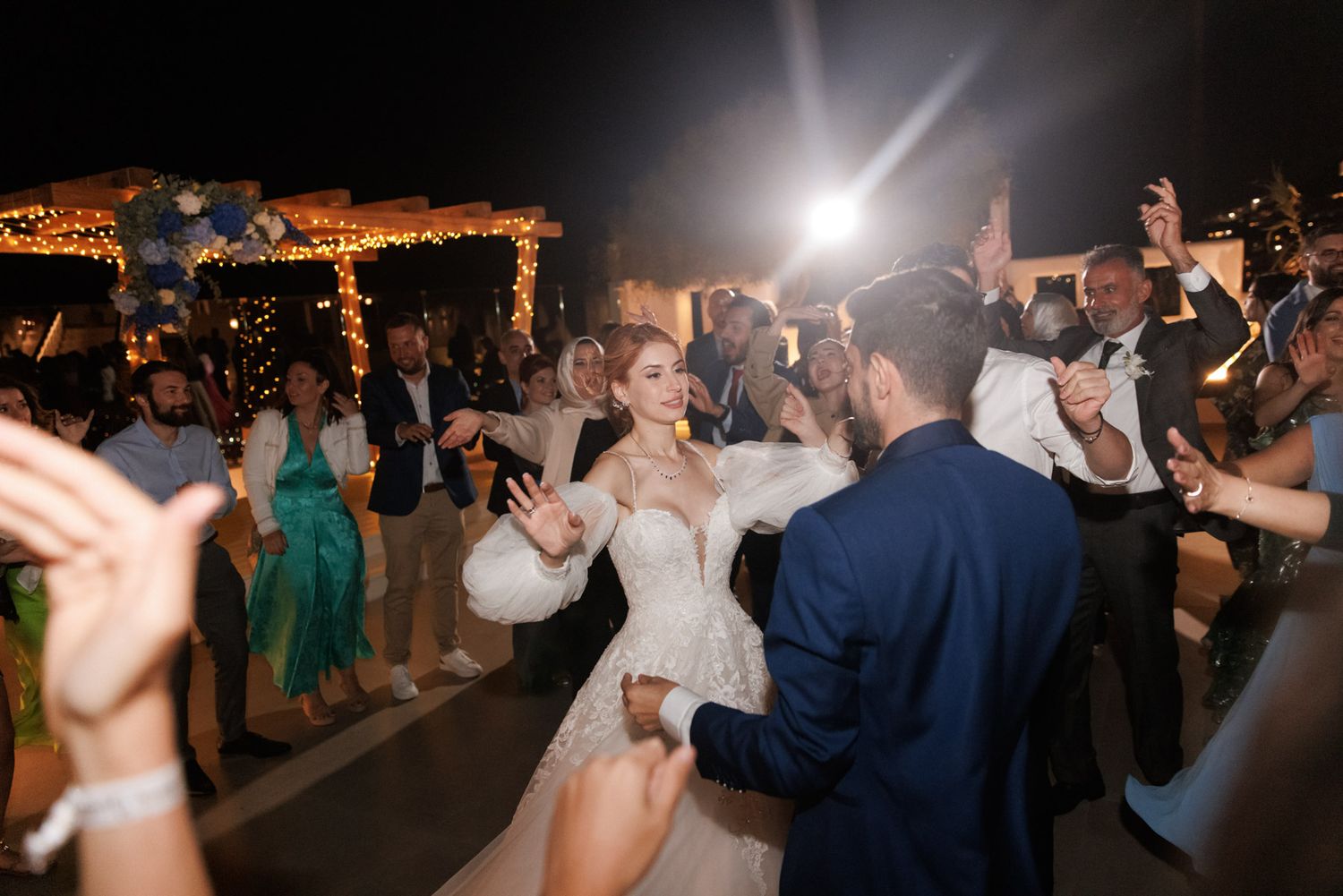Bride in lace gown and groom in navy suit dancing joyfully surrounded by guests at outdoor wedding reception.