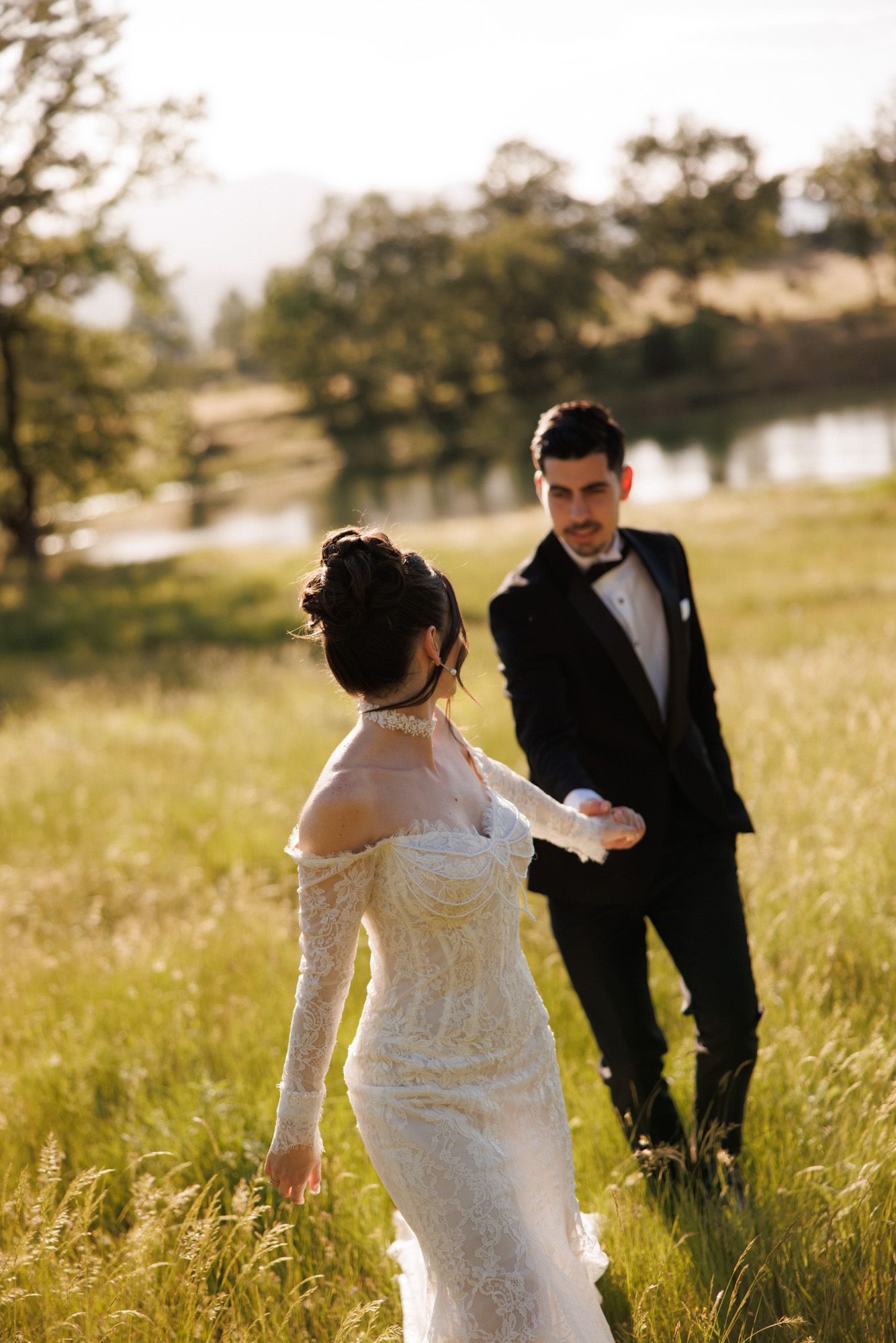 Bride in lace gown holds groom's hand in sunlit meadow, with trees and lake in background.
