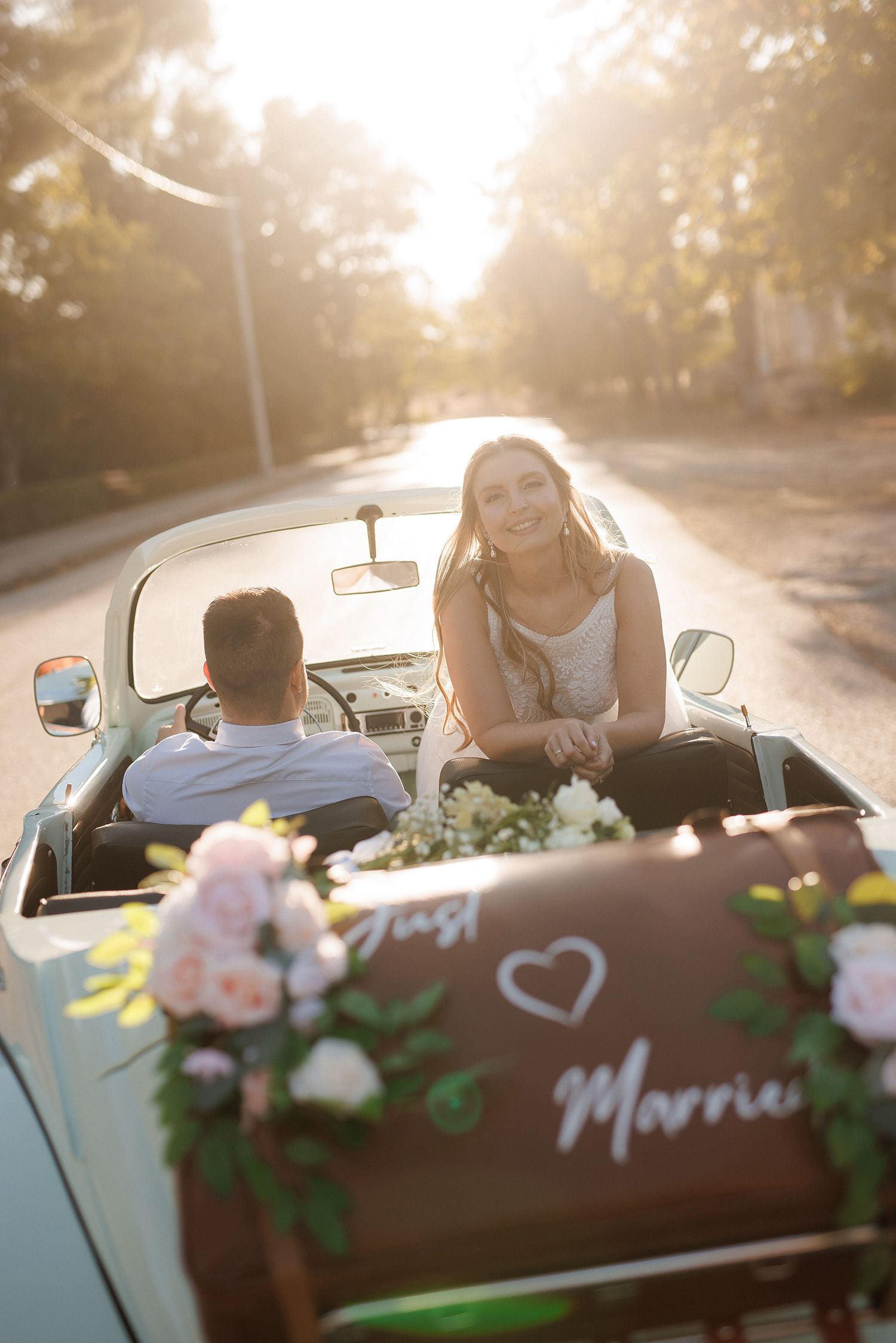 Newlywed couple riding in a vintage convertible decorated with flowers and a Just Married sign in golden sunlight.