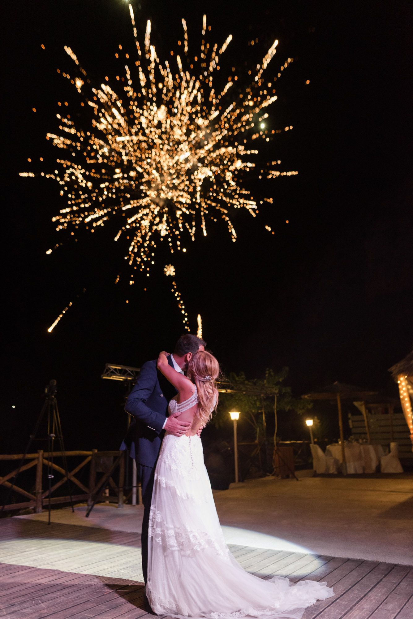 Newlyweds share a romantic kiss under spectacular fireworks display at their outdoor wedding reception.