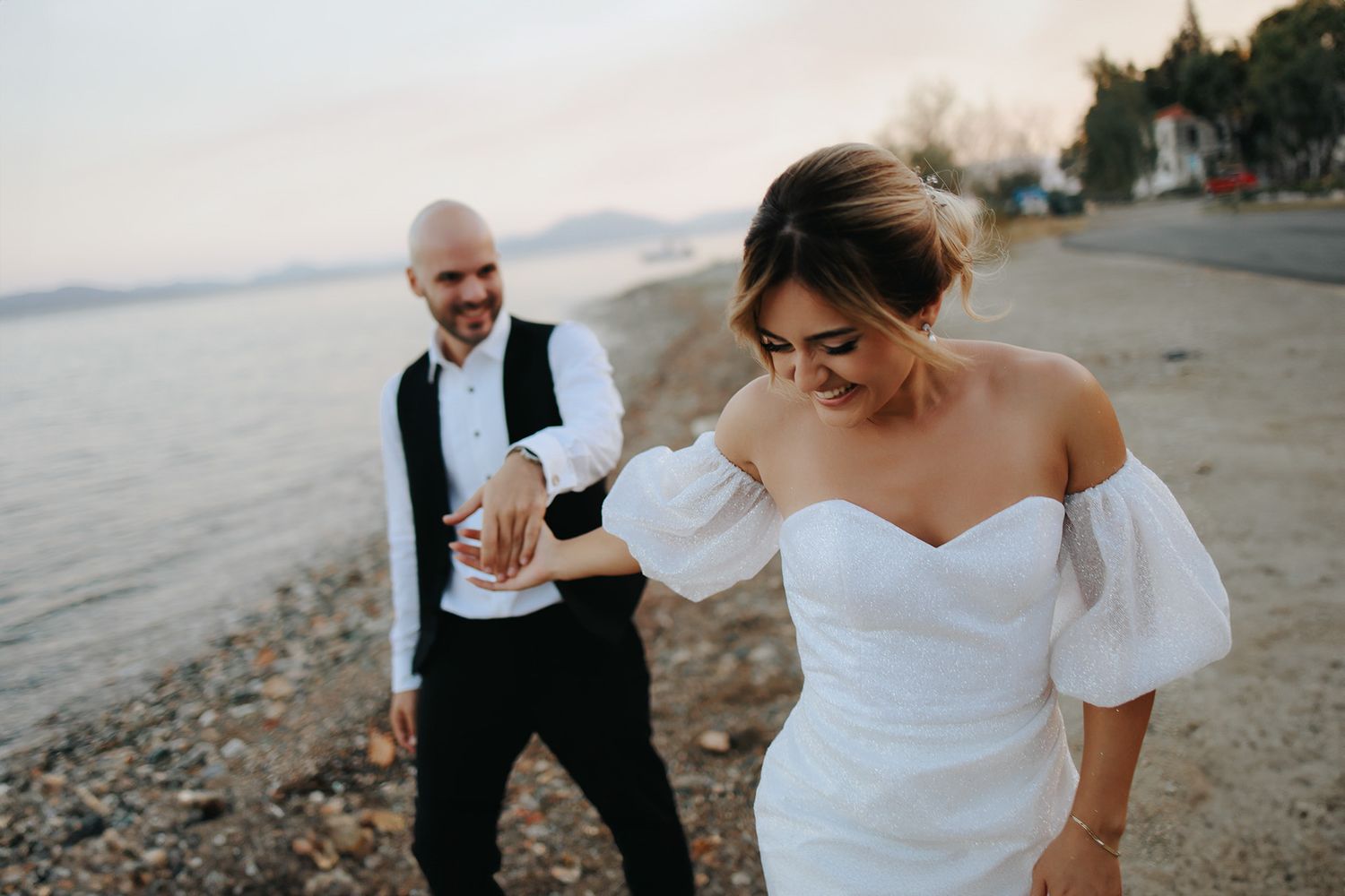 Couple walking along a rocky beach at sunset with bride wearing an off-shoulder white dress.