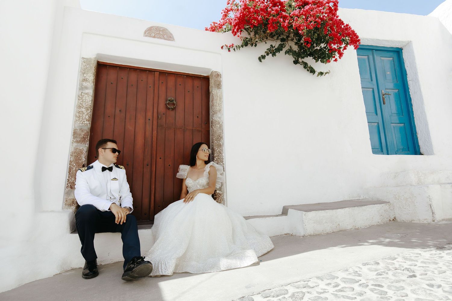 Bride and groom sitting by a rustic wooden door with white walls and red flowers in a Mediterranean setting.