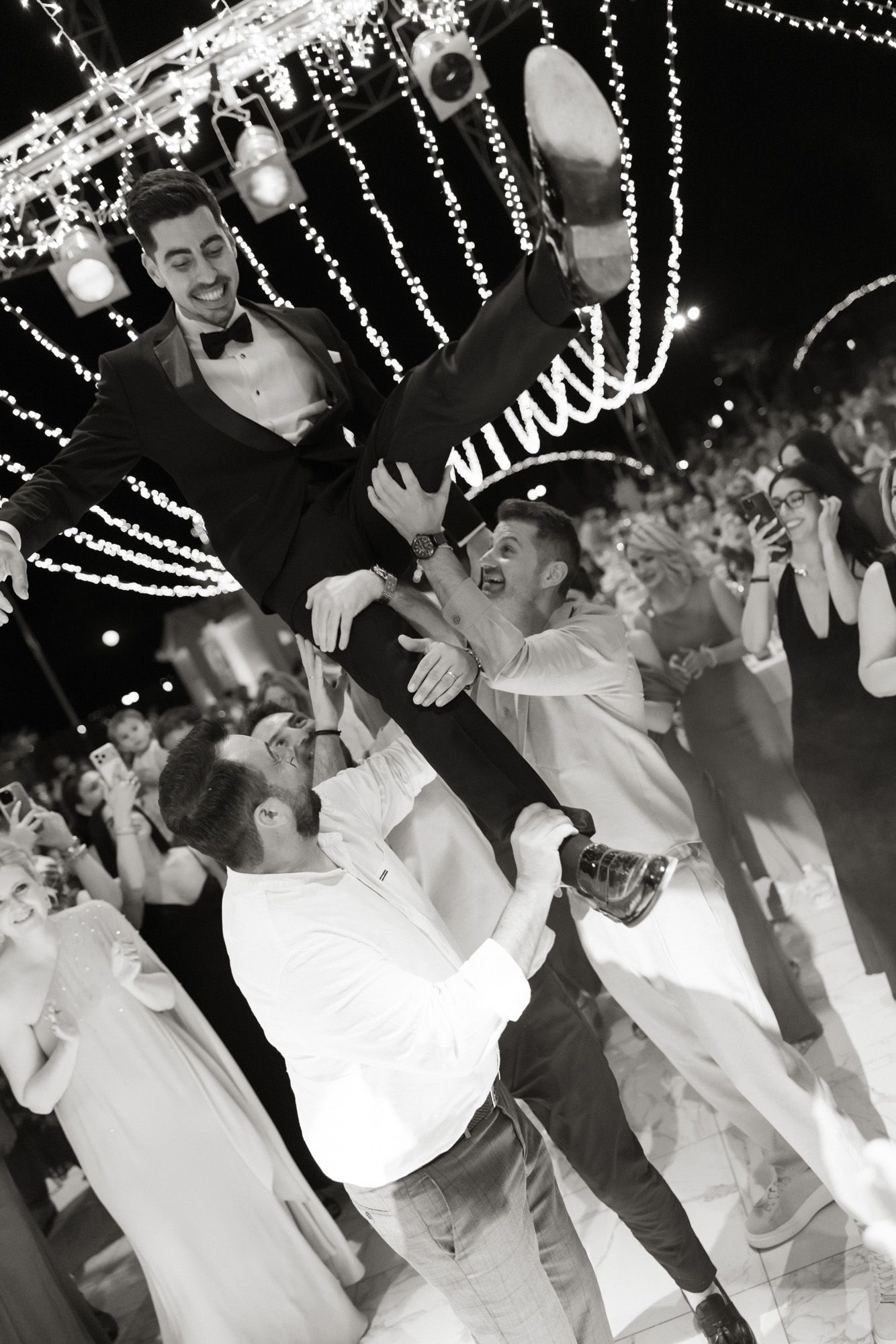 Black and white photo of groom being lifted by groomsmen on dance floor at outdoor wedding reception.