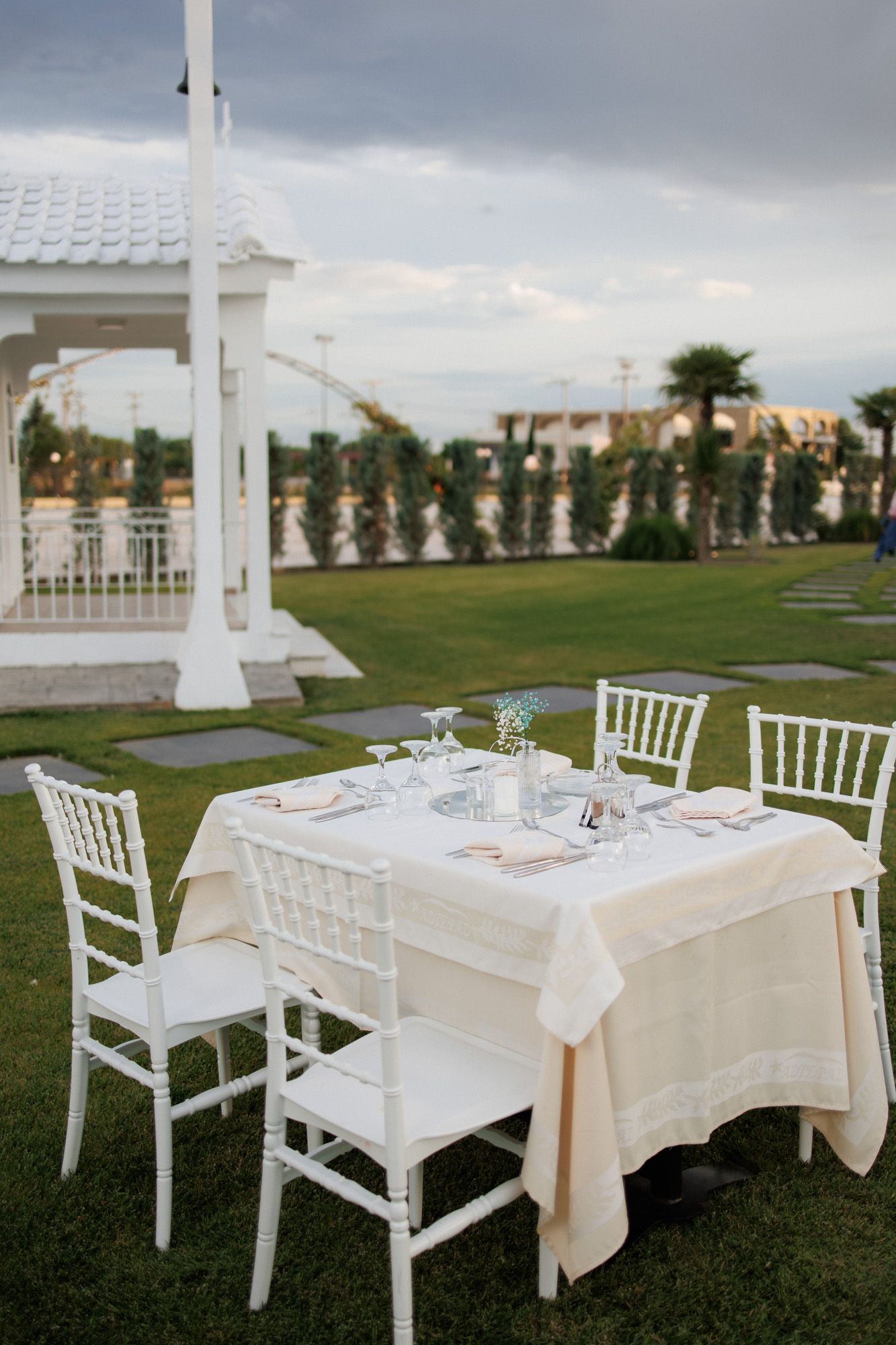 Elegant outdoor dining table with white Chiavari chairs and cream linens set on green lawn near a gazebo.