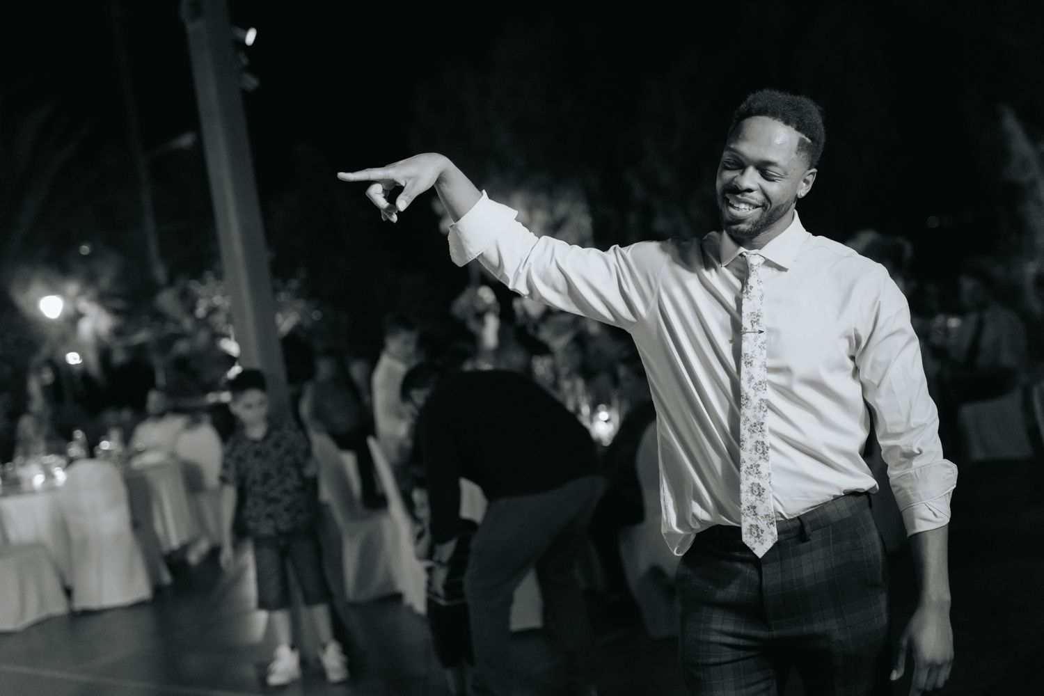 Black and white photo of a smiling man in a dress shirt and tie pointing at a wedding reception dance floor.