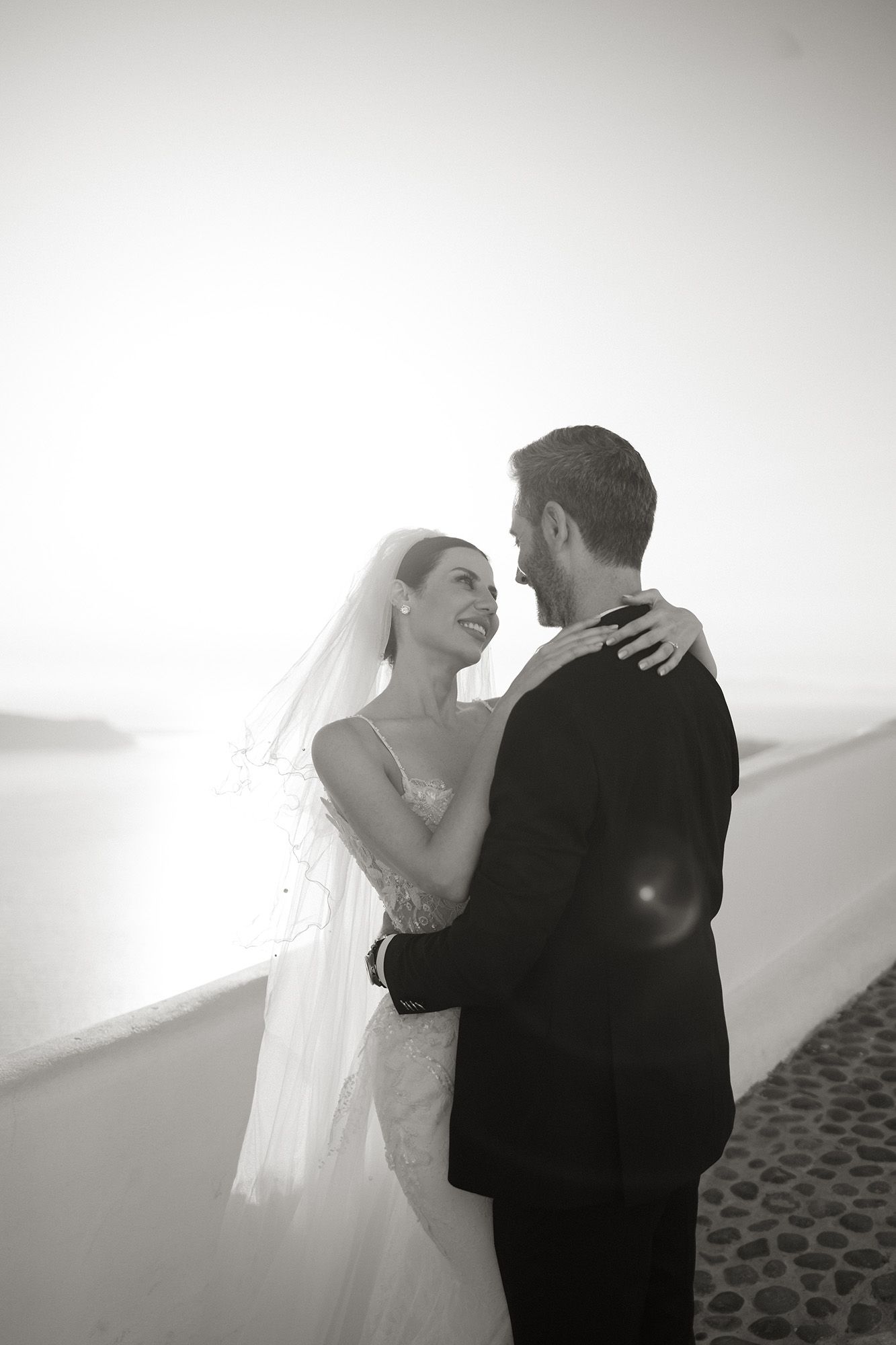 Black and white photo of a smiling bride and groom embracing near the sea on their wedding day.
