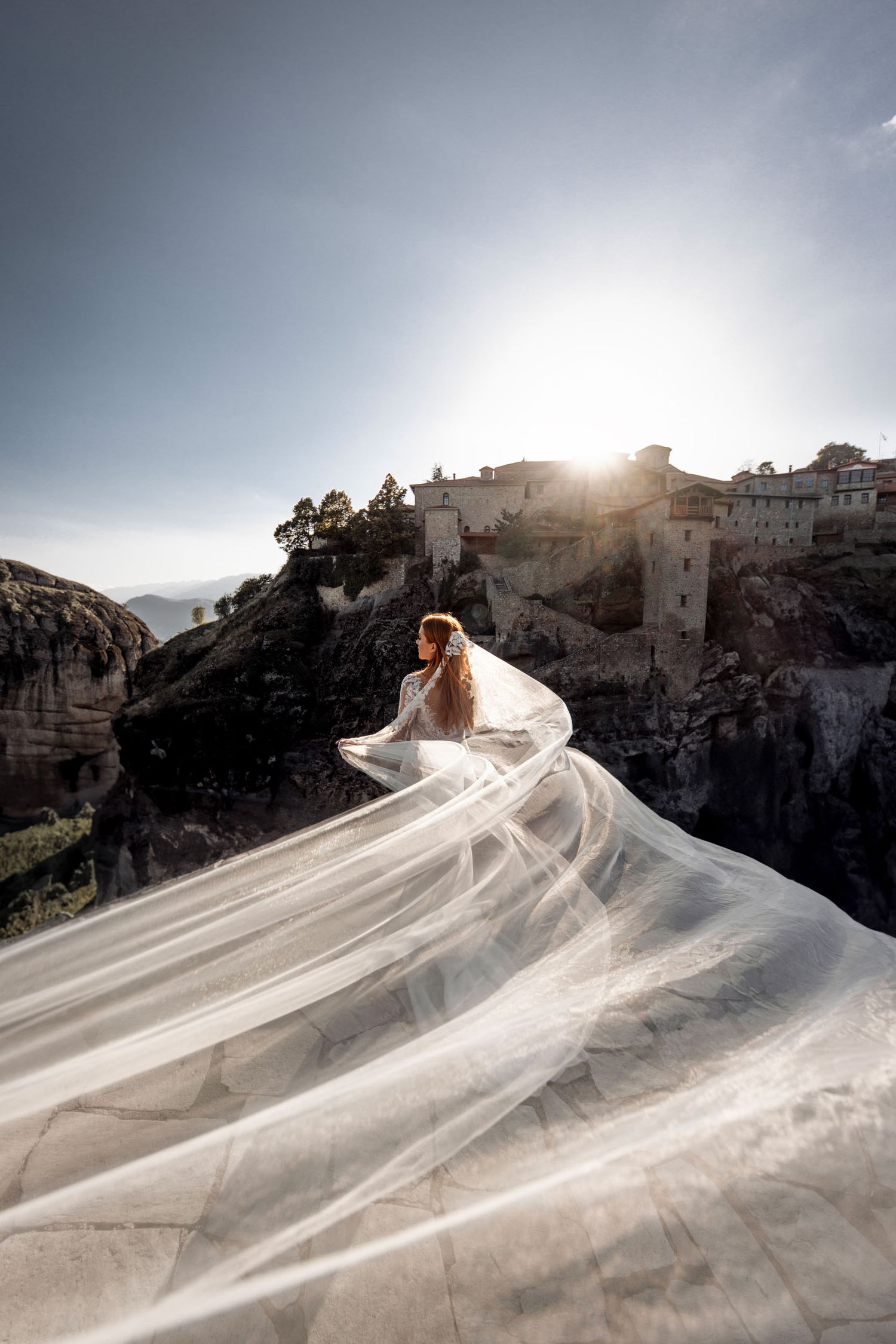Flowing white wedding dress and veil dramatically billowing in the wind against a cliffside mountain backdrop at sunset.