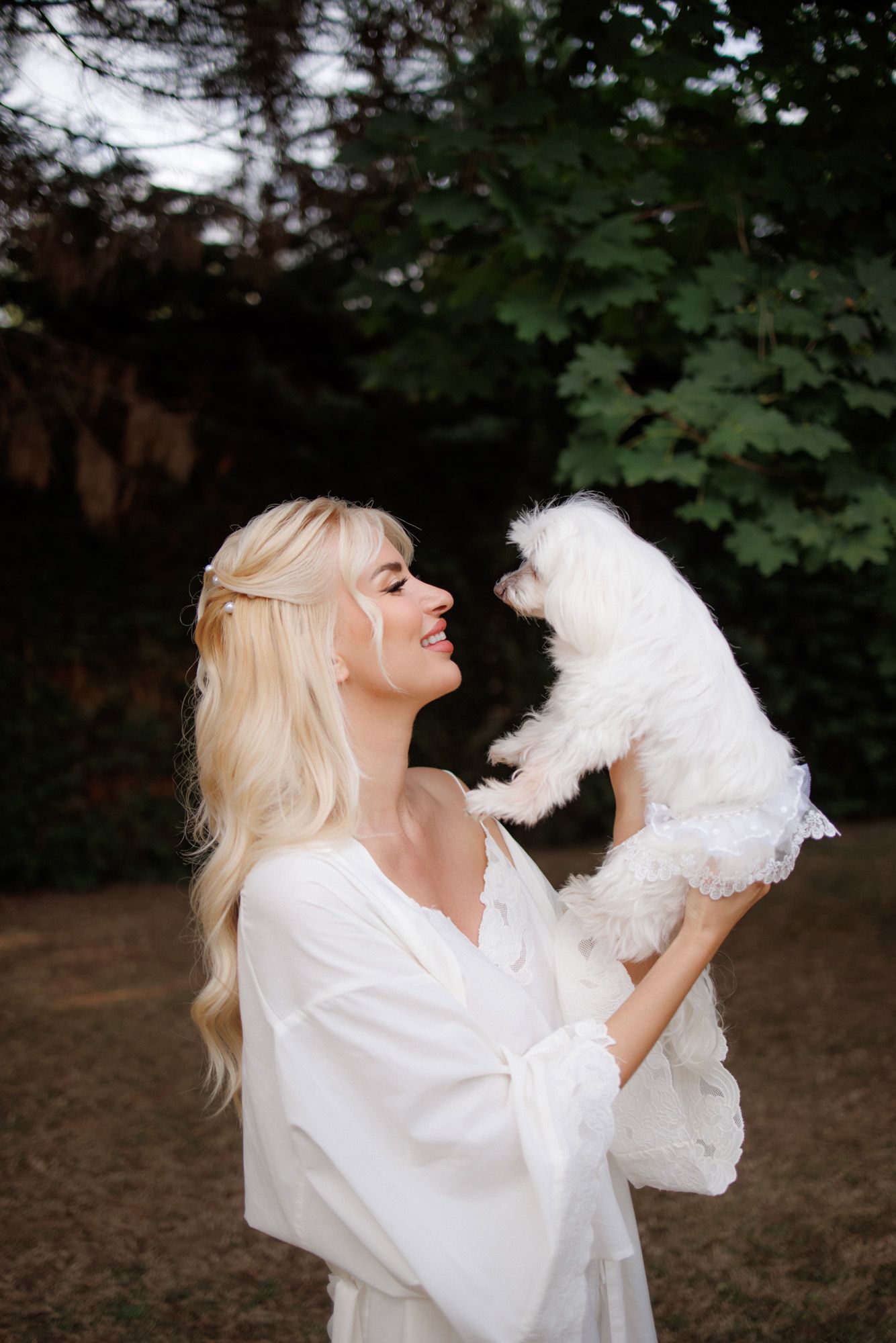 Joyful blonde woman in white robe lifting a fluffy white dog outdoors among lush green trees.