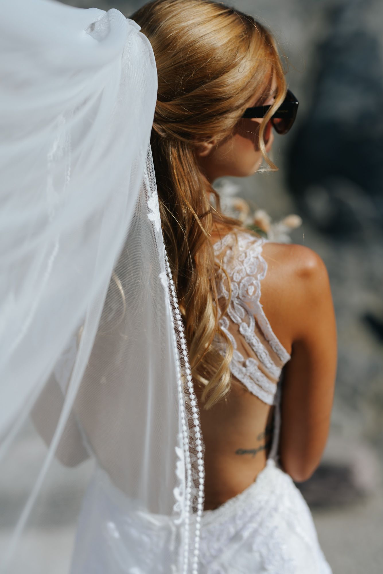 Bride in backless lace wedding gown with pearl-trimmed veil and sunglasses poses at rocky location.
