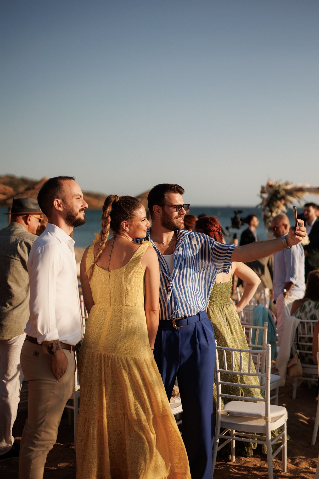 Three wedding guests pose for a selfie at a beachside ceremony during golden hour.
