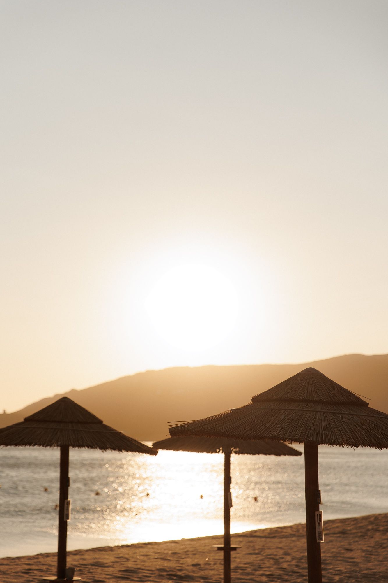 Three rustic thatched beach umbrellas silhouetted against a golden sunset over calm ocean waters.