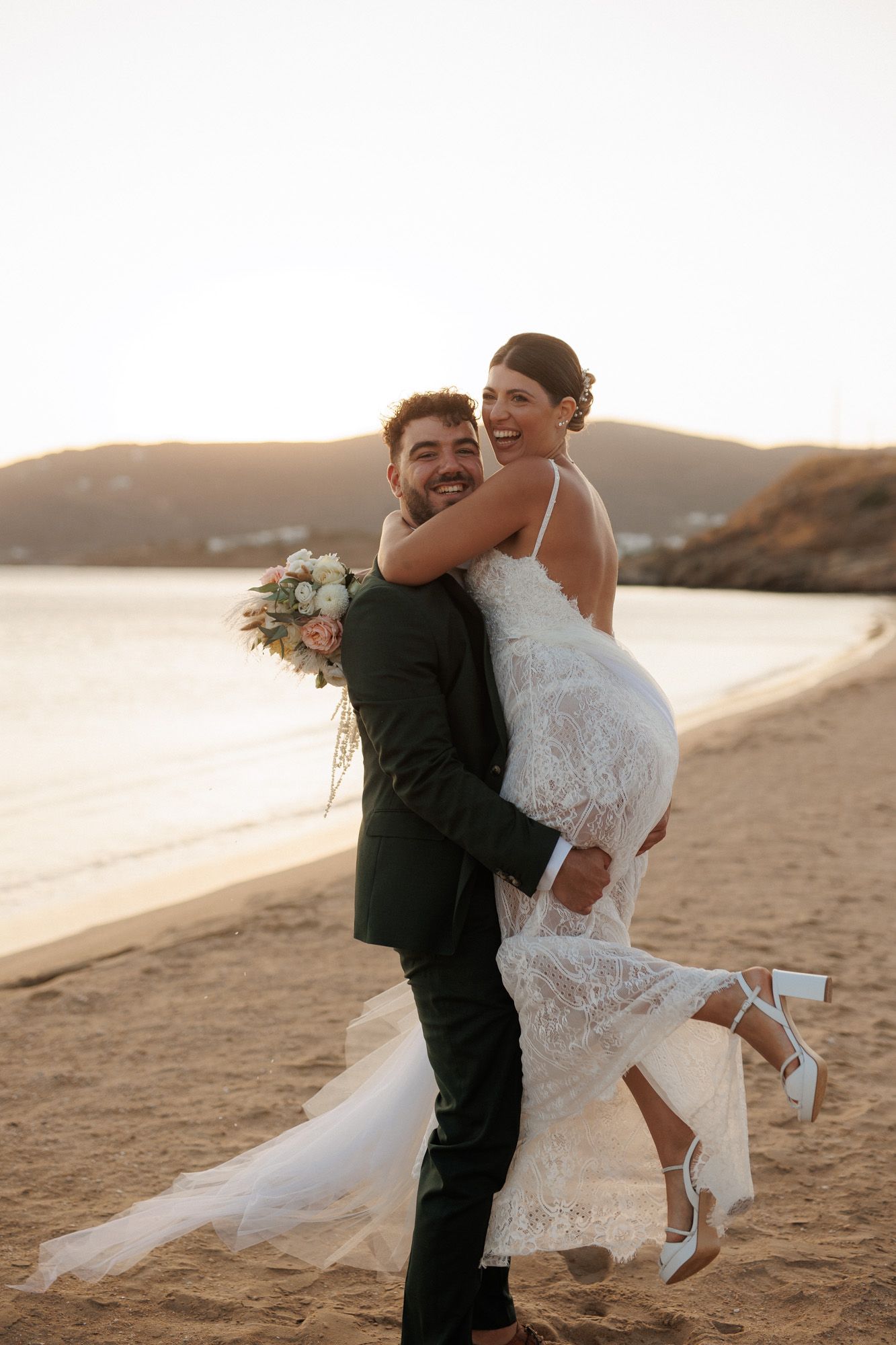 Groom lifts laughing bride on golden sandy beach at sunset during wedding celebration.