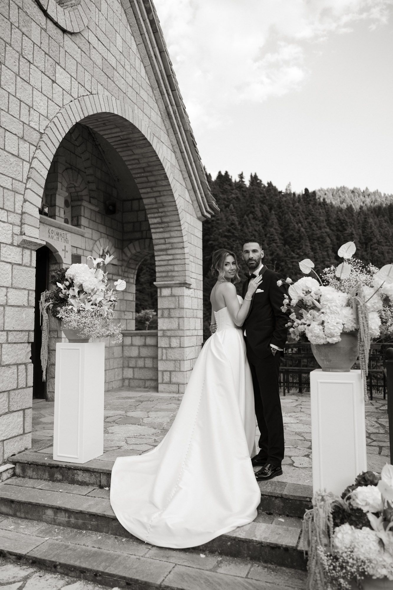 Bride and groom pose on stone church steps flanked by floral arrangements in black and white photo.
