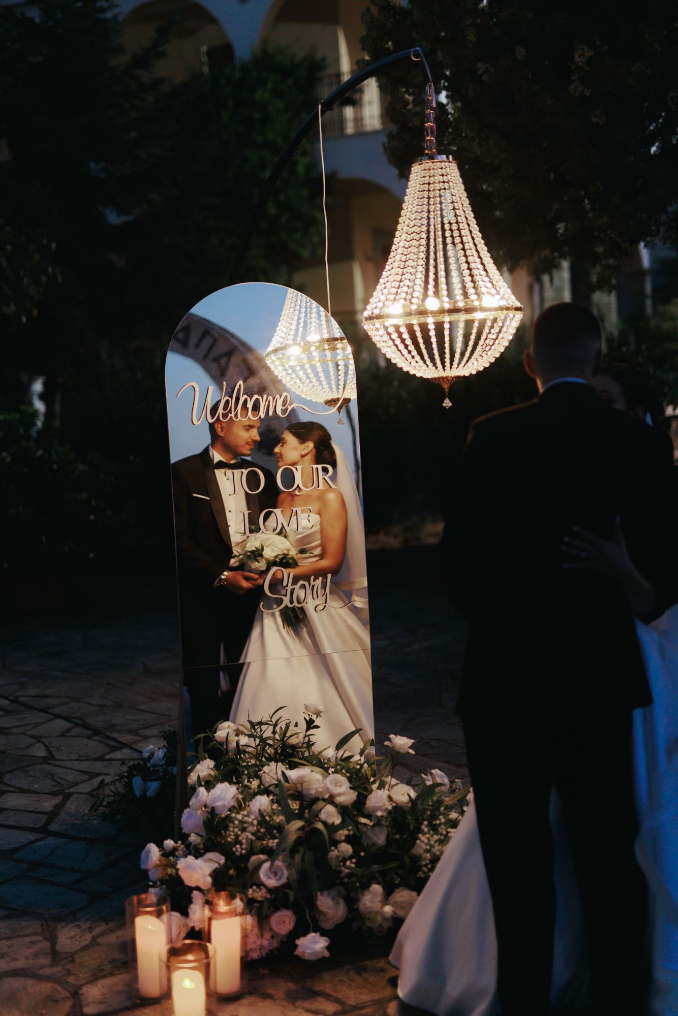 Elegant wedding mirror sign with couple photo, chandelier, candles, and floral arrangement at outdoor evening reception.