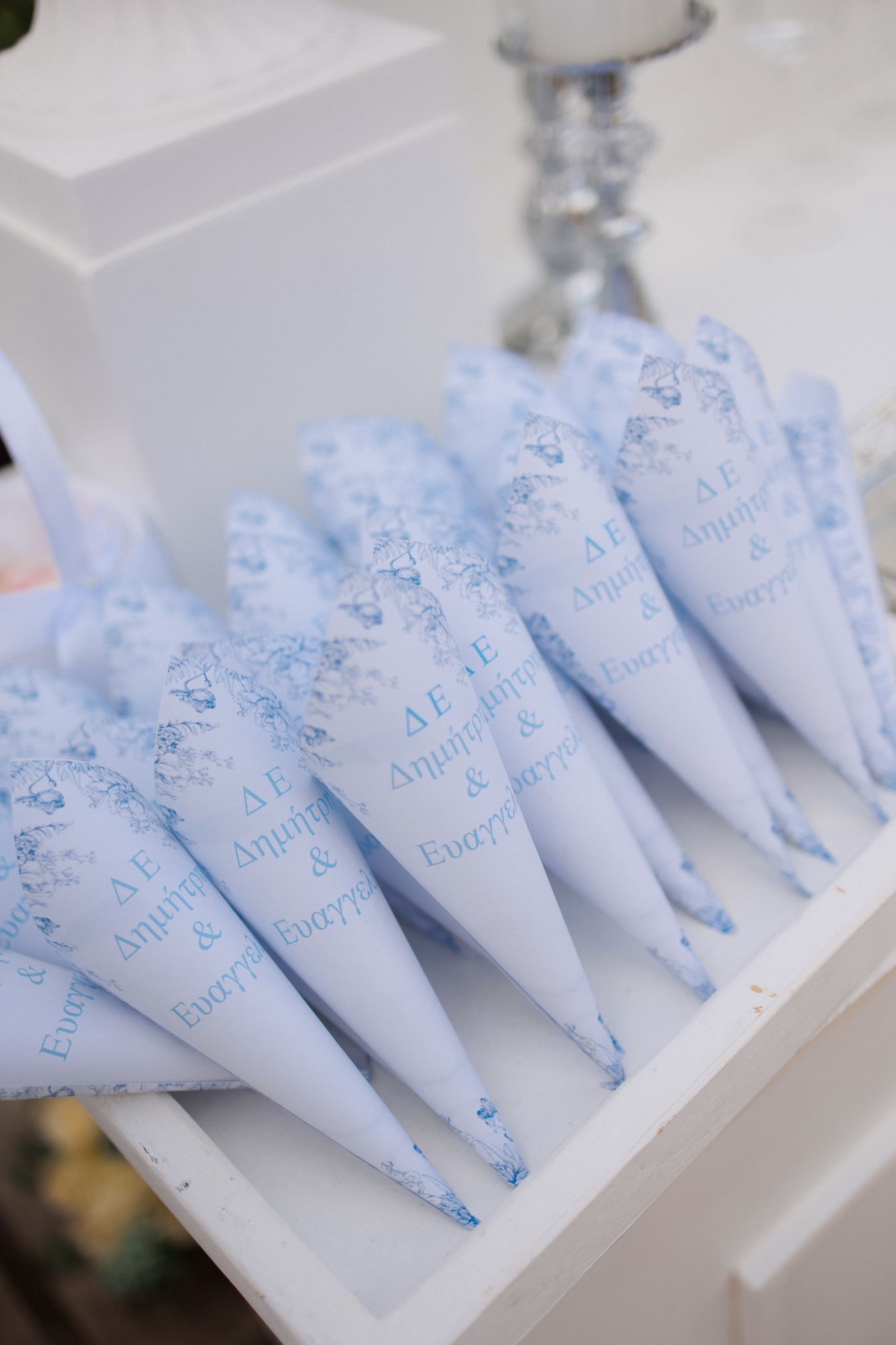 White paper cones with blue Greek text and floral design displayed on a white shelf for a wedding celebration.