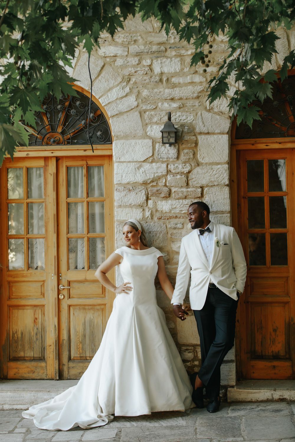 Bride in white gown and groom in white tuxedo jacket hold hands before rustic stone building with wooden doors.