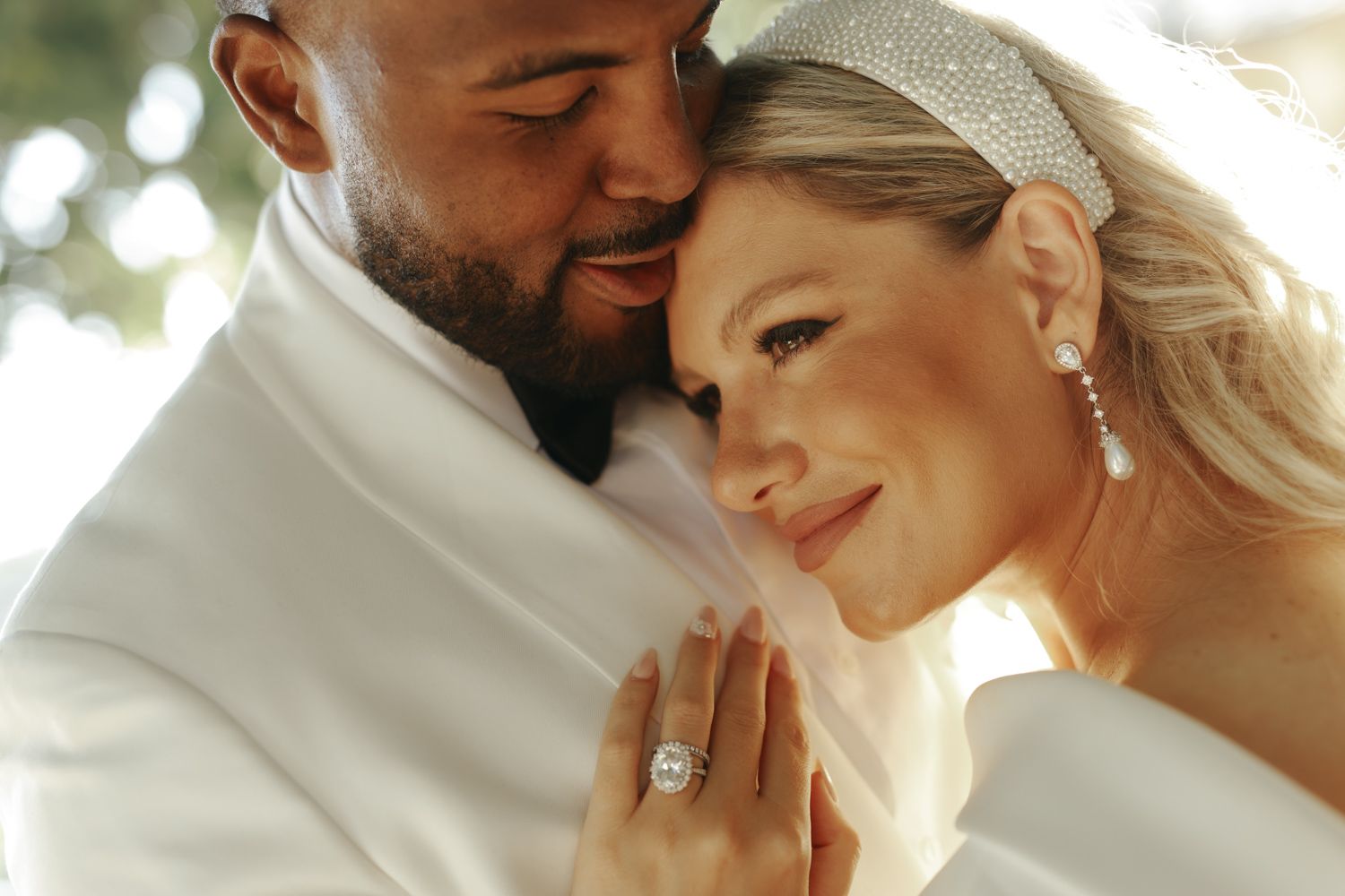 Bride and groom in white wedding attire share a romantic close moment, showcasing a stunning diamond engagement ring.