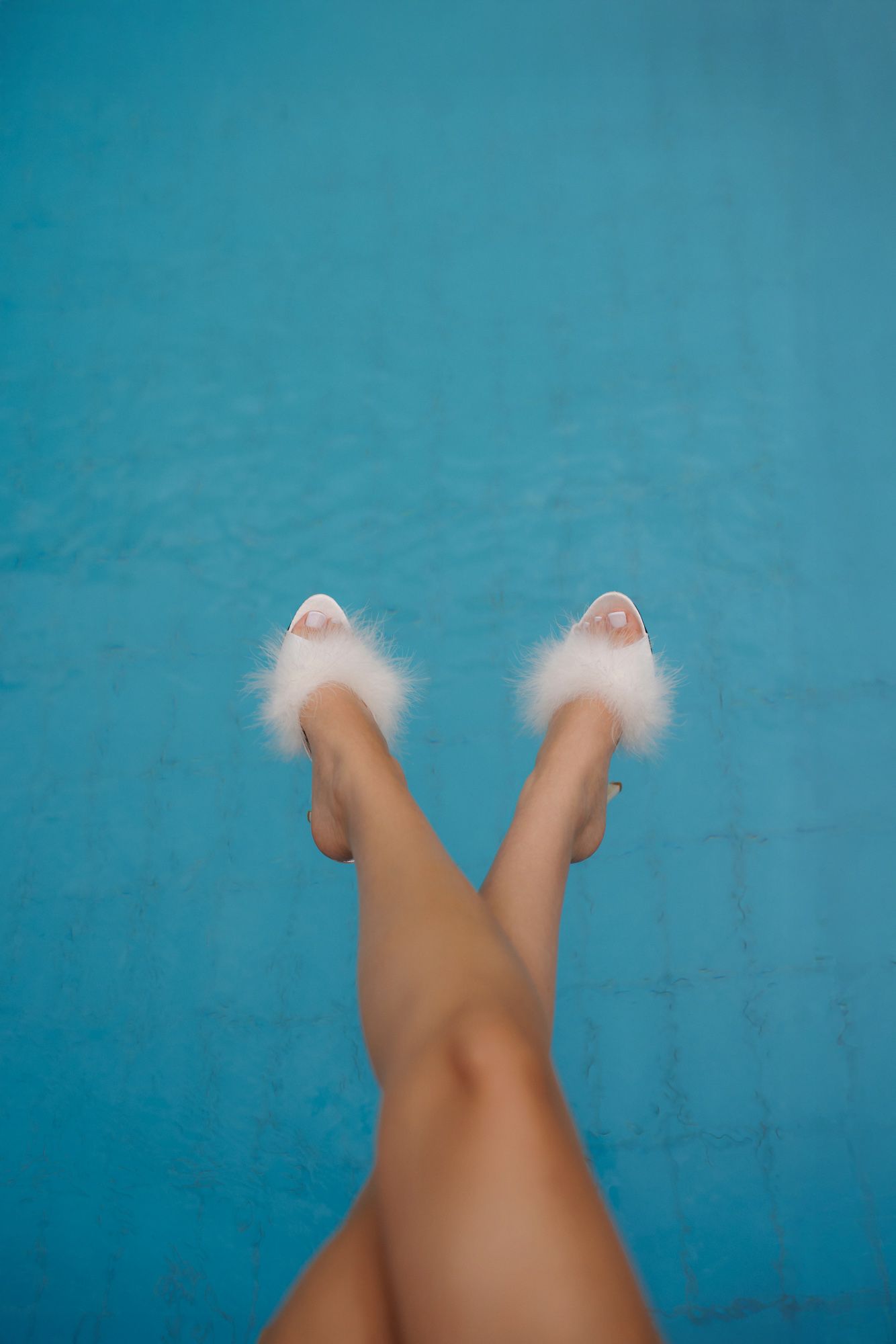 Womans legs raised in the air wearing white fluffy feather mule heels over a blue swimming pool.