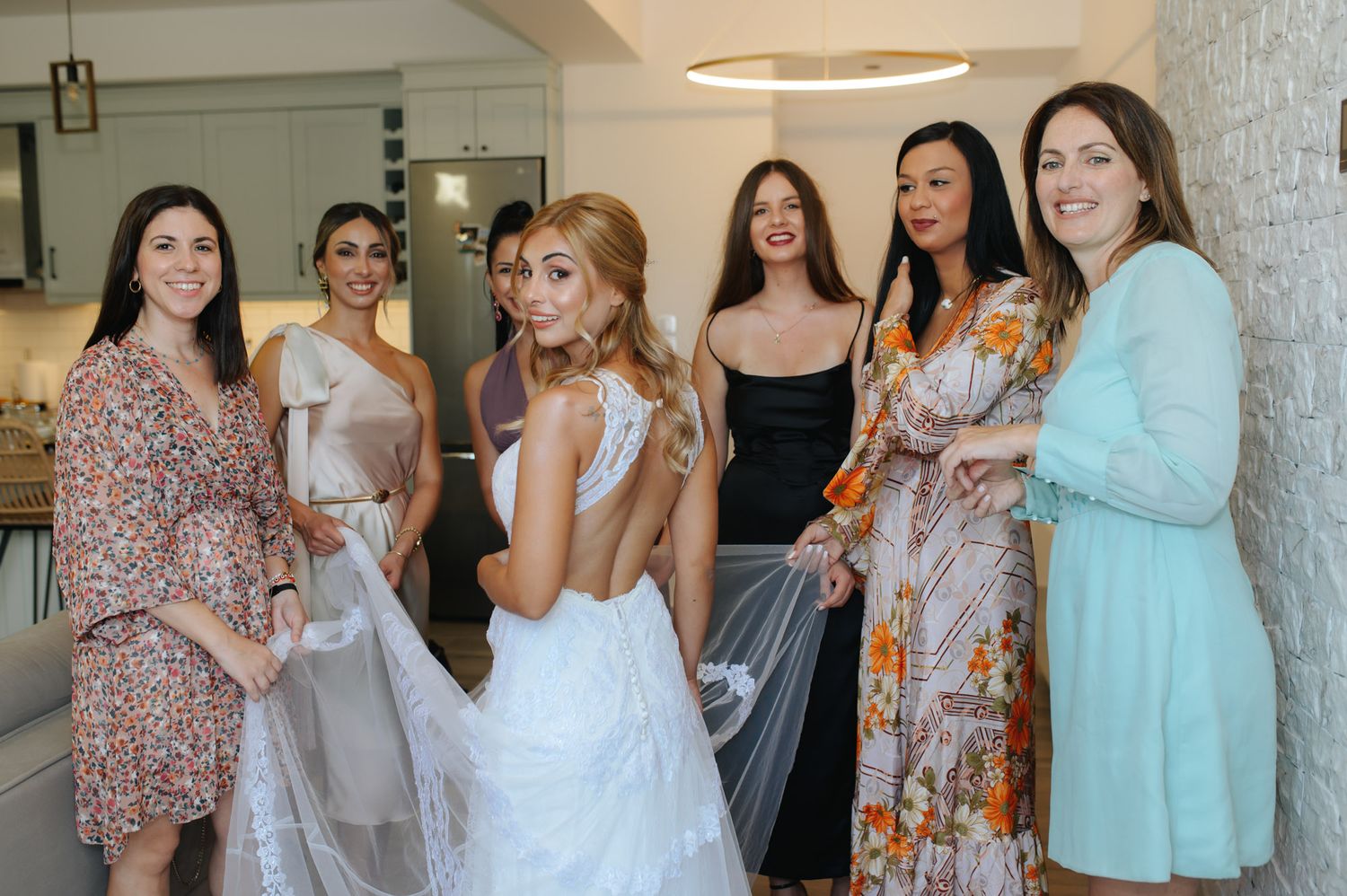 Bride in elegant backless white wedding gown posing with five bridesmaids in colorful dresses indoors.