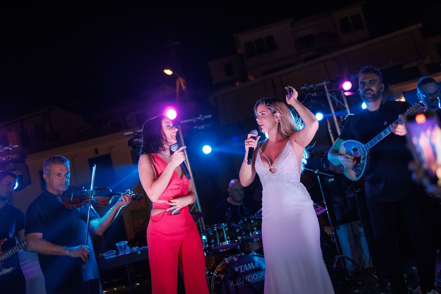 Two women singing on stage with a live band performing at an outdoor night event.