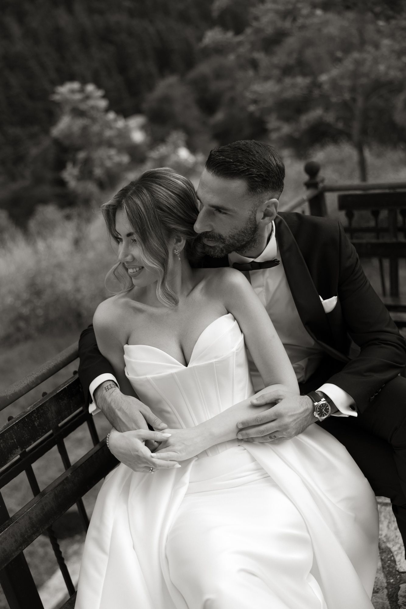 Groom tenderly embraces smiling bride on bench with lush green landscape in black and white portrait.