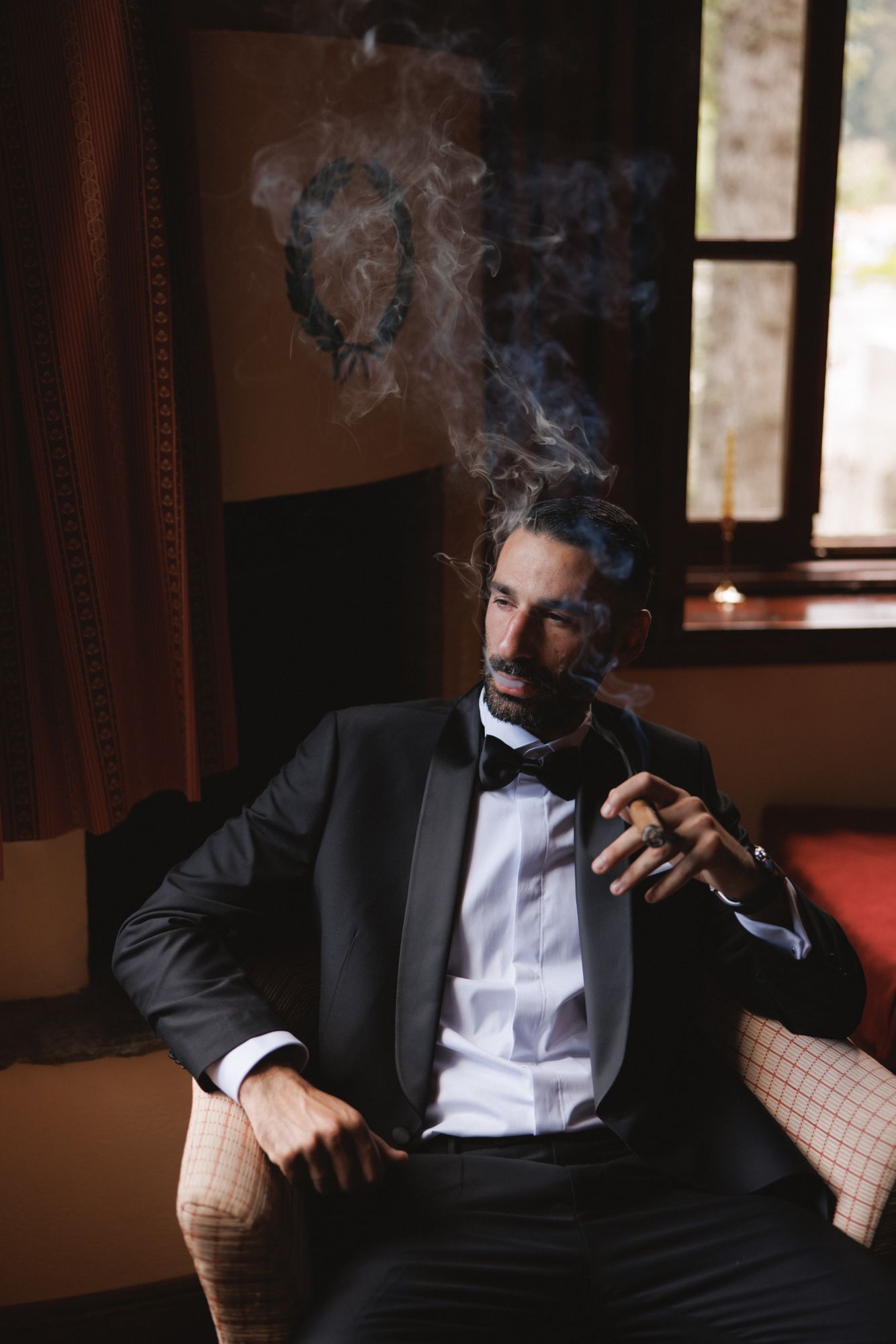 Sophisticated man in black tuxedo with bow tie smoking a cigar while seated in a luxury interior.