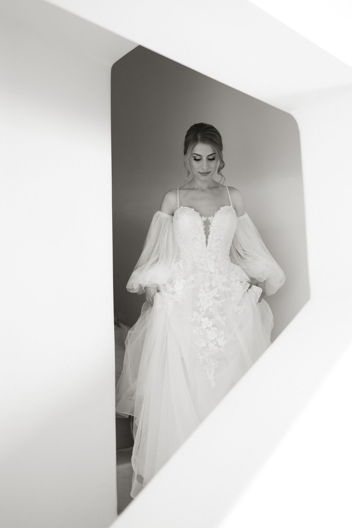 Black and white portrait of a bride in a strapless lace ball gown framed in a geometric white architectural niche.