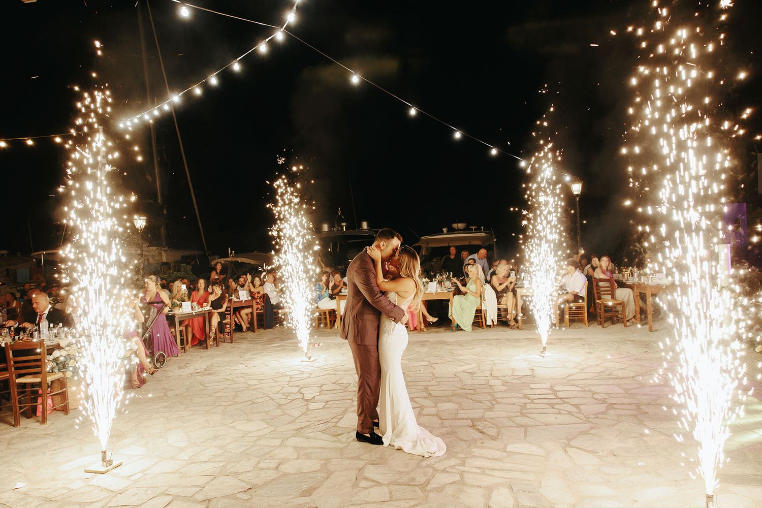 A couple shares their first dance at a nighttime wedding reception surrounded by sparkling firework fountains.