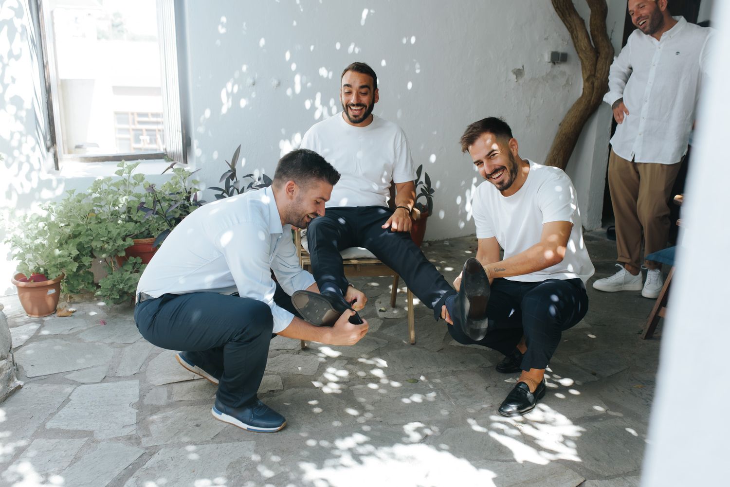 Three groomsmen in white shirts laughing and celebrating together in a sunlit courtyard.