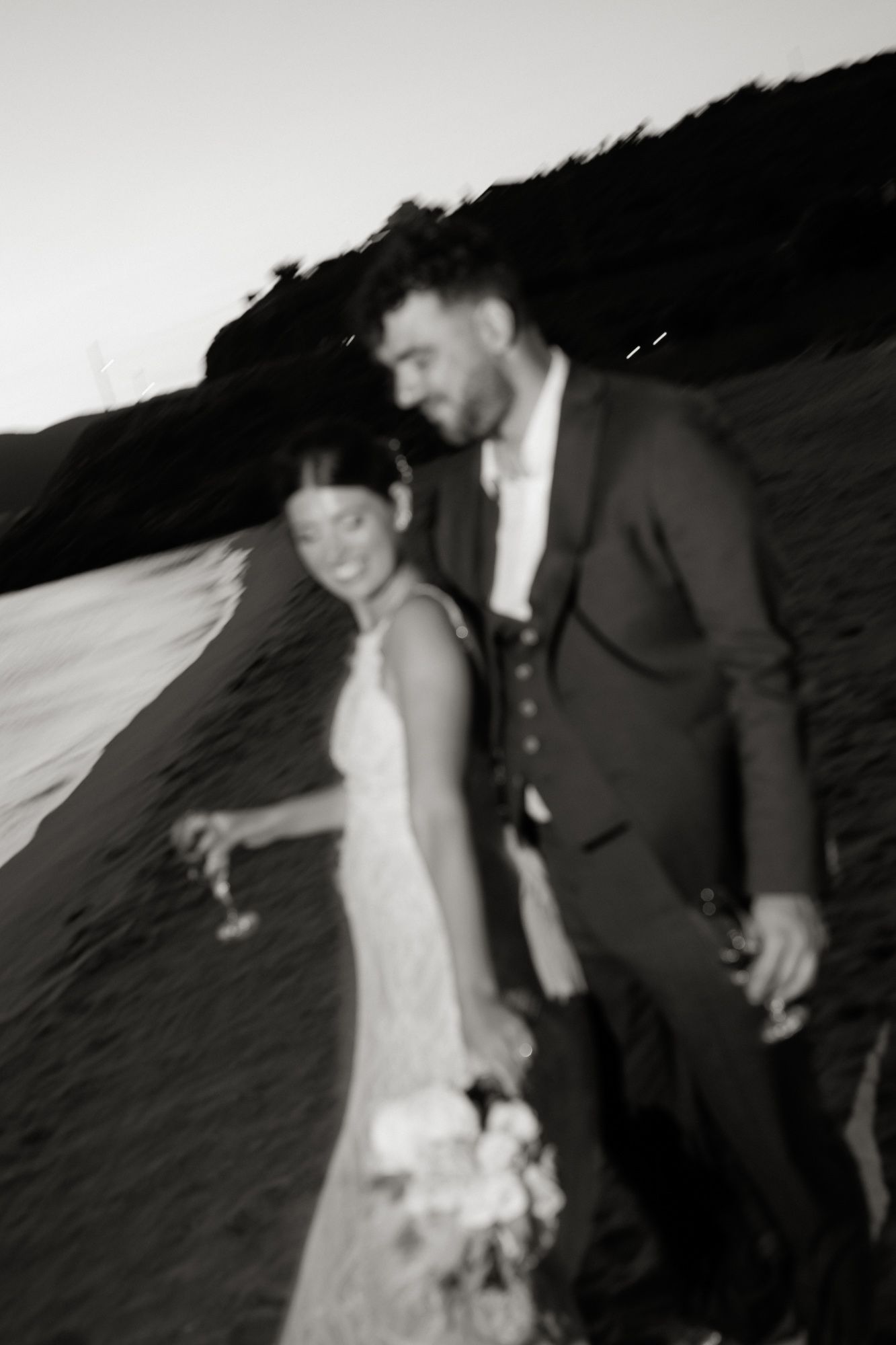 Black and white photo of bride and groom walking hand in hand along beach at dusk.