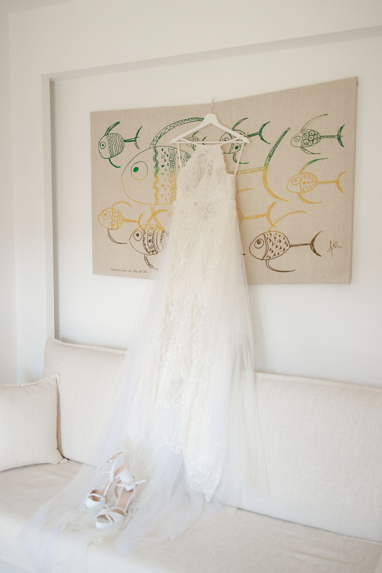 Elegant lace wedding dress hanging on hanger above white sofa with bridal heels below.