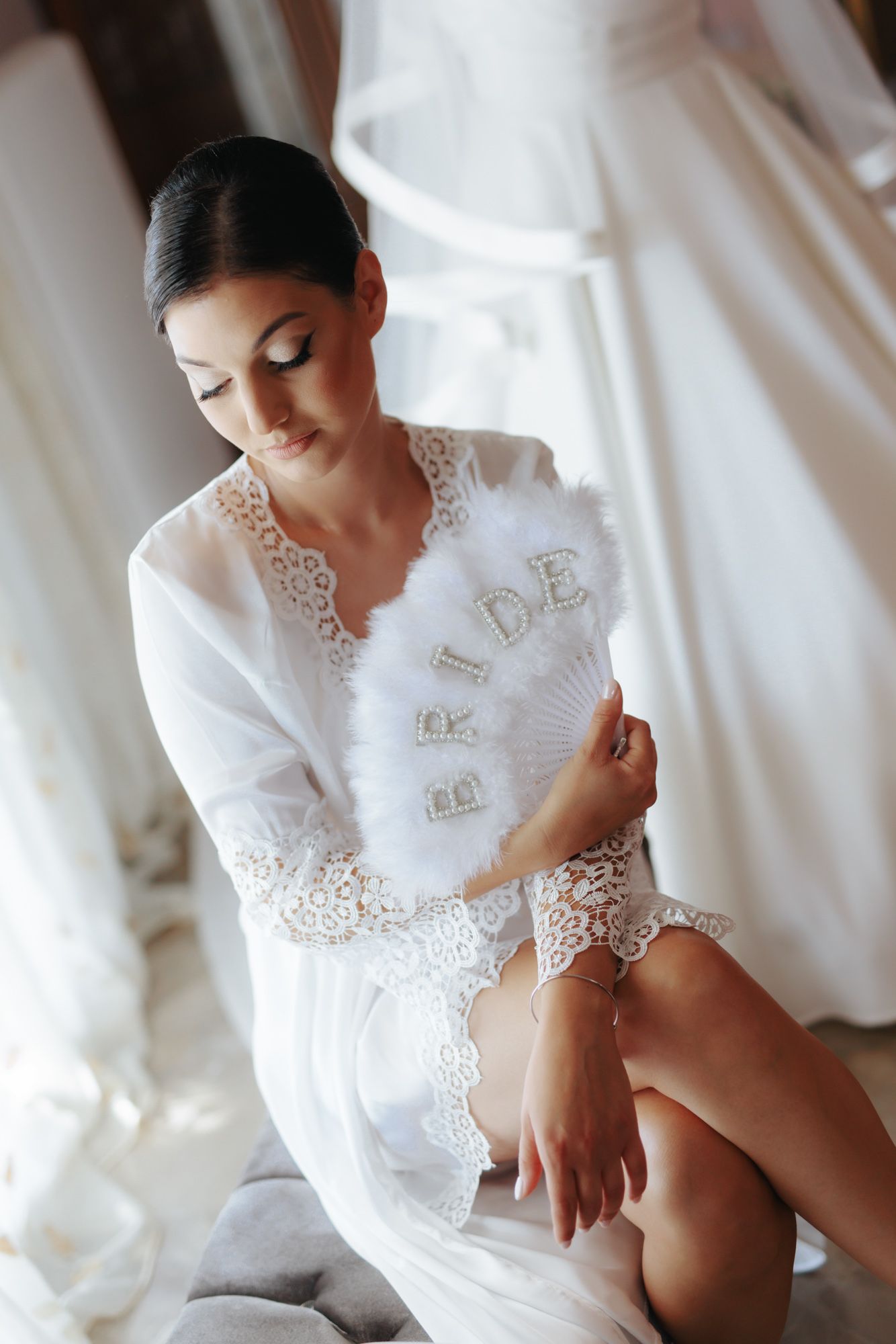 Bride in white lace robe holding a feather fan with BRIDE letters, sitting elegantly in a bridal boutique.