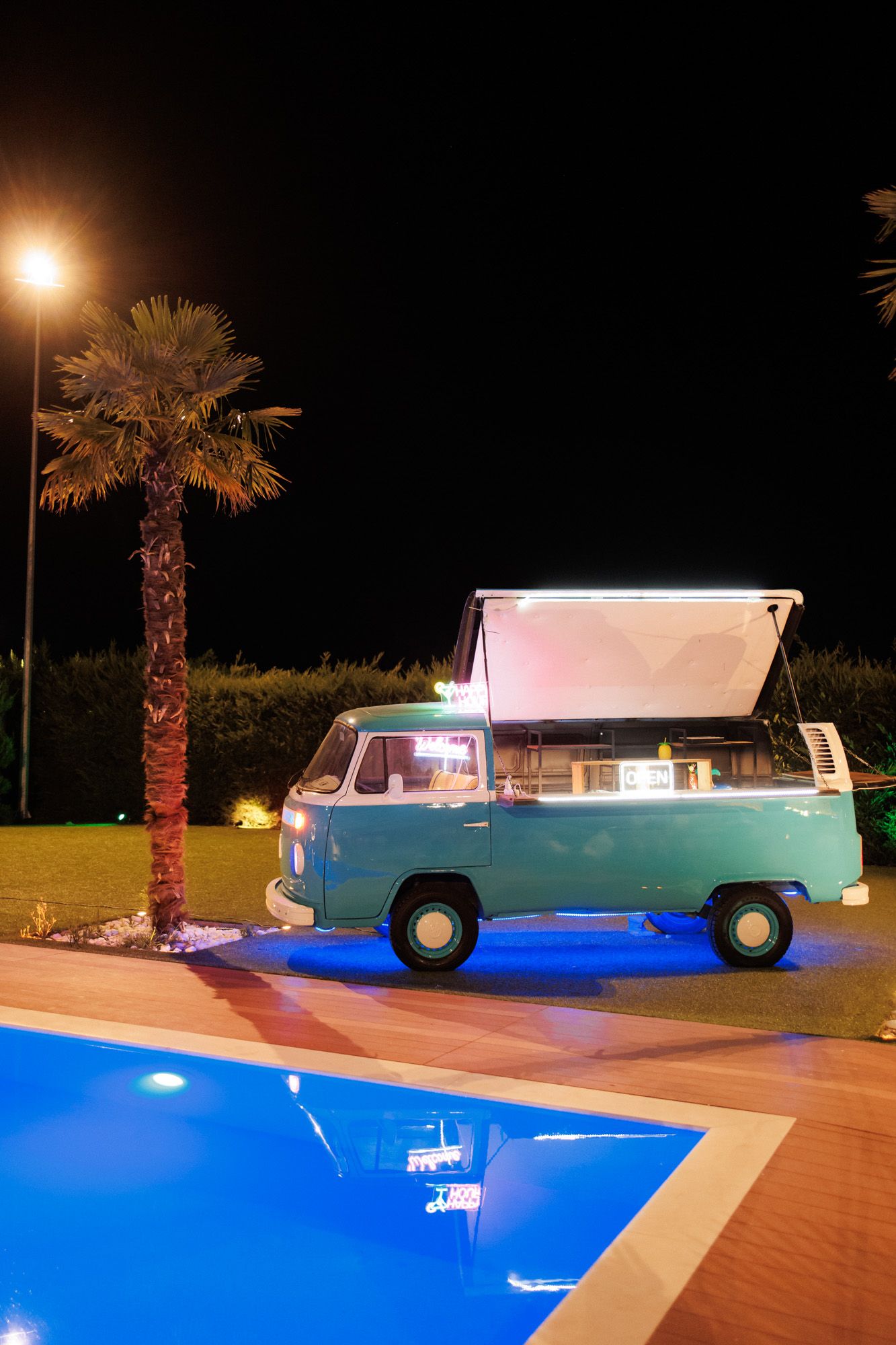 Vintage teal VW camper van converted to food truck parked beside illuminated pool and palm tree at night.