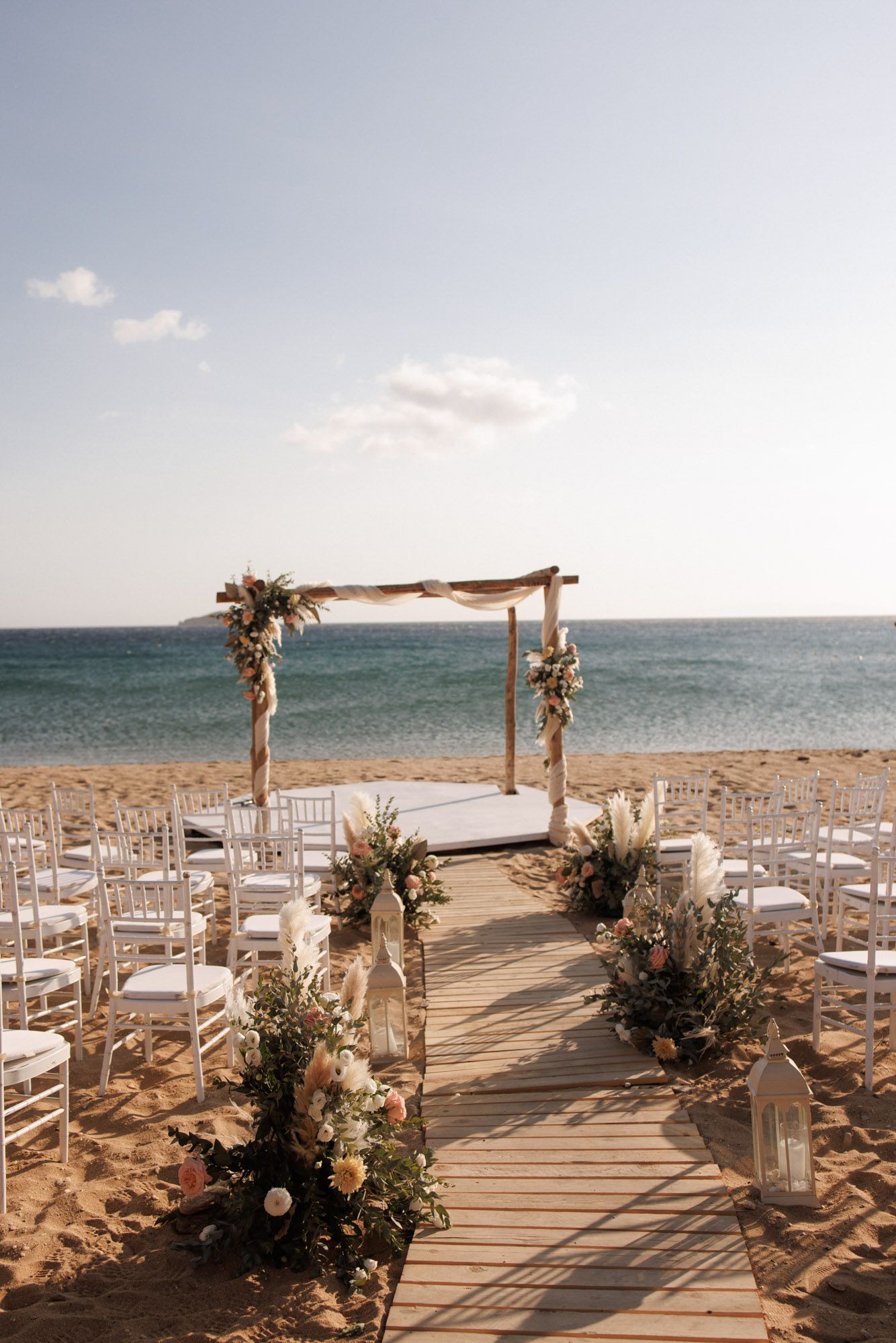 Elegant beach wedding aisle with wooden arch, white chairs, and ocean backdrop.