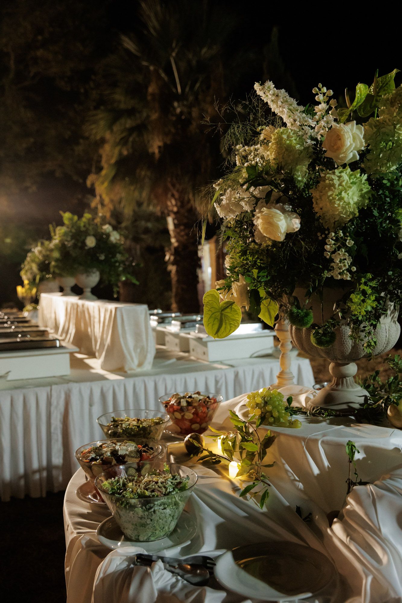 Elegant outdoor wedding reception buffet table adorned with tall floral arrangements of white roses and greenery at night.