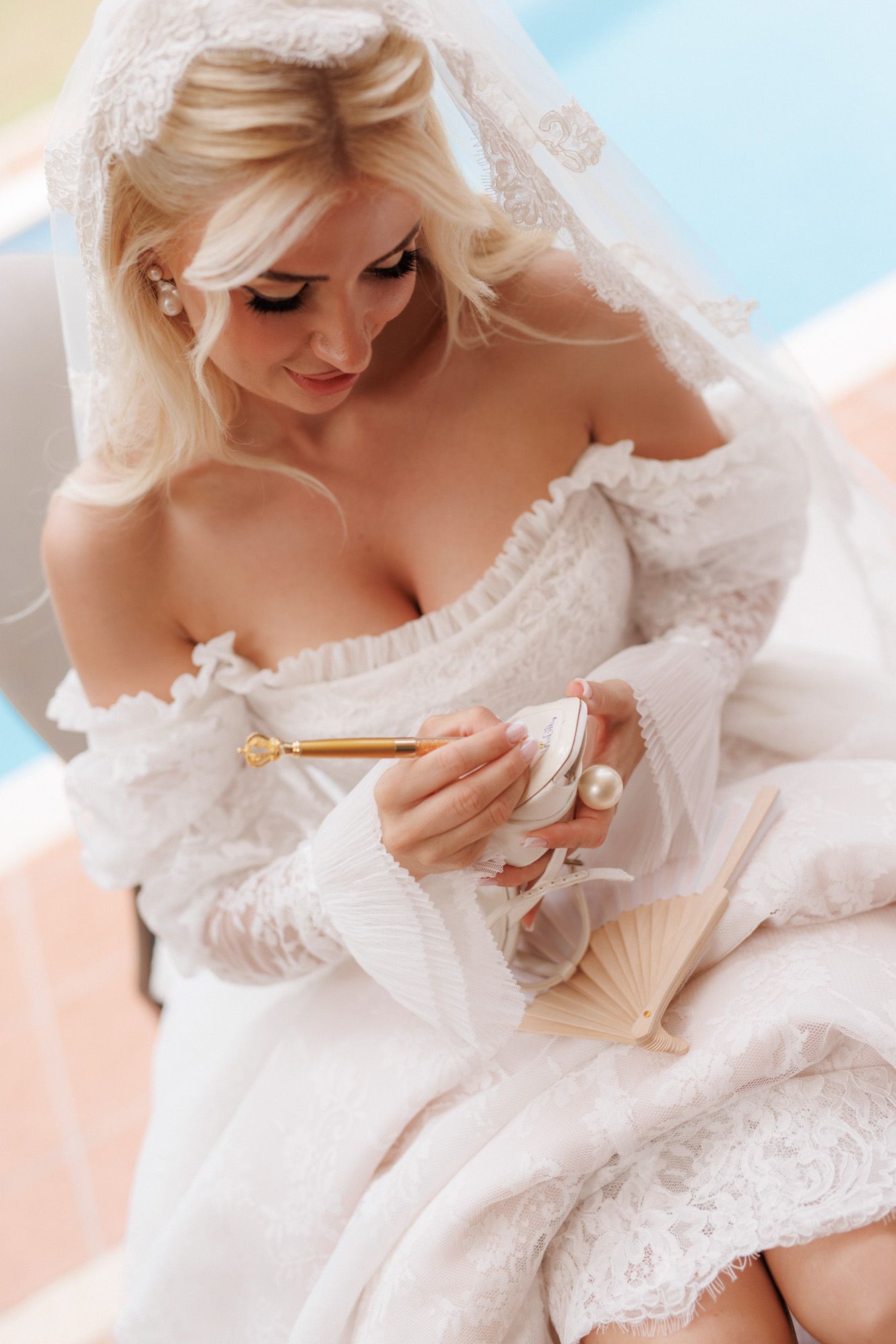 Elegant bride in white lace off-shoulder gown holding a fan and mirror, wearing a delicate veil.