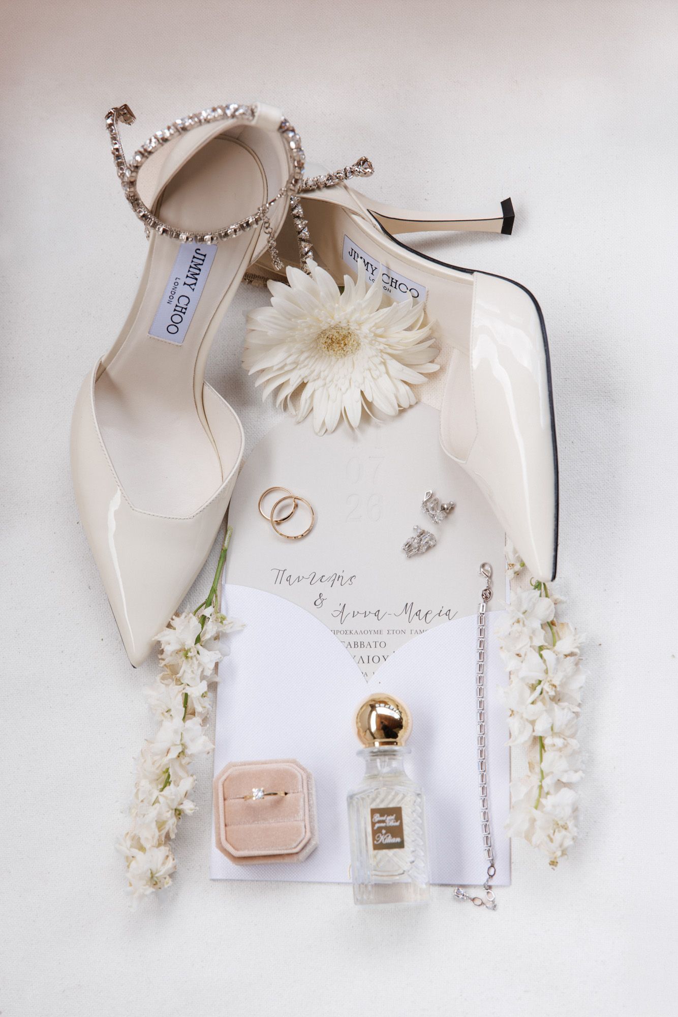 Elegant bridal flat lay with Jimmy Choo heels, wedding rings, perfume, and floral invitation suite.