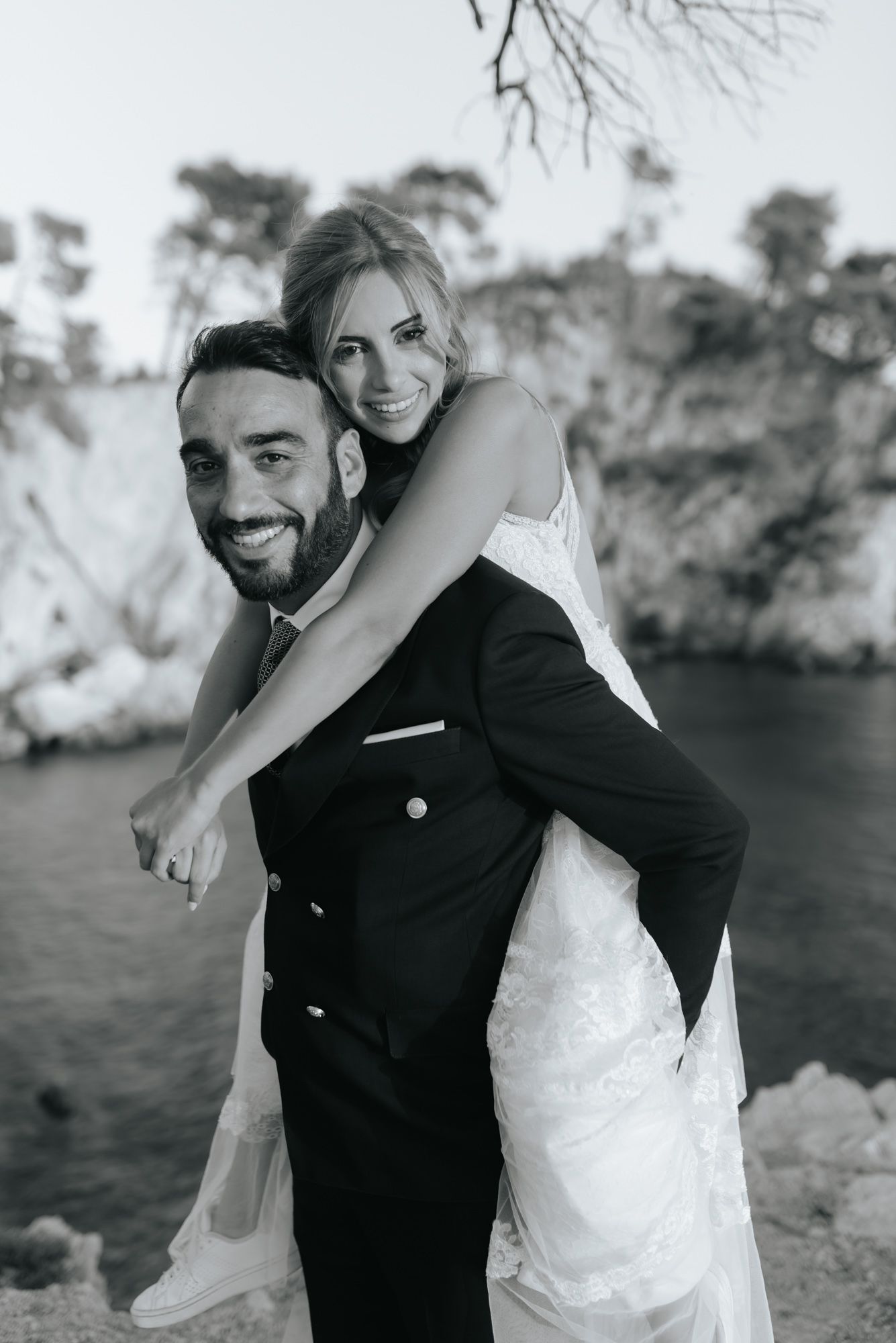 Black and white photo of smiling groom giving bride a piggyback ride near rocky coastal cliffs.