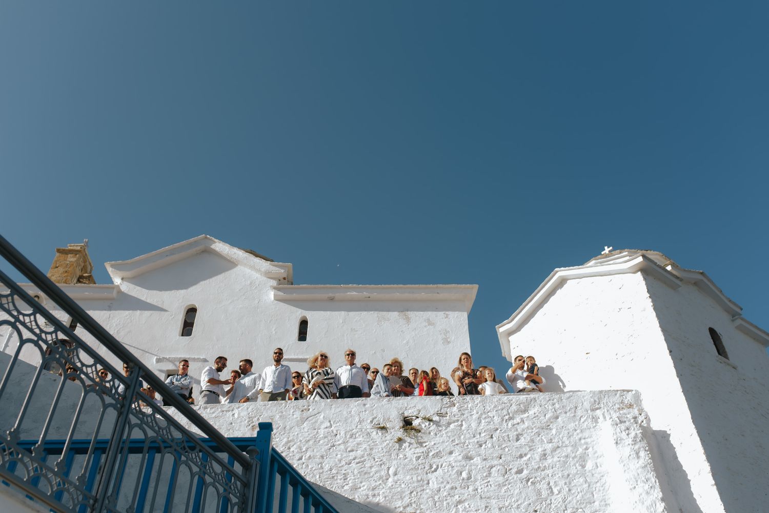 Wedding guests gathered on a white-walled Greek church terrace under a bright blue summer sky.