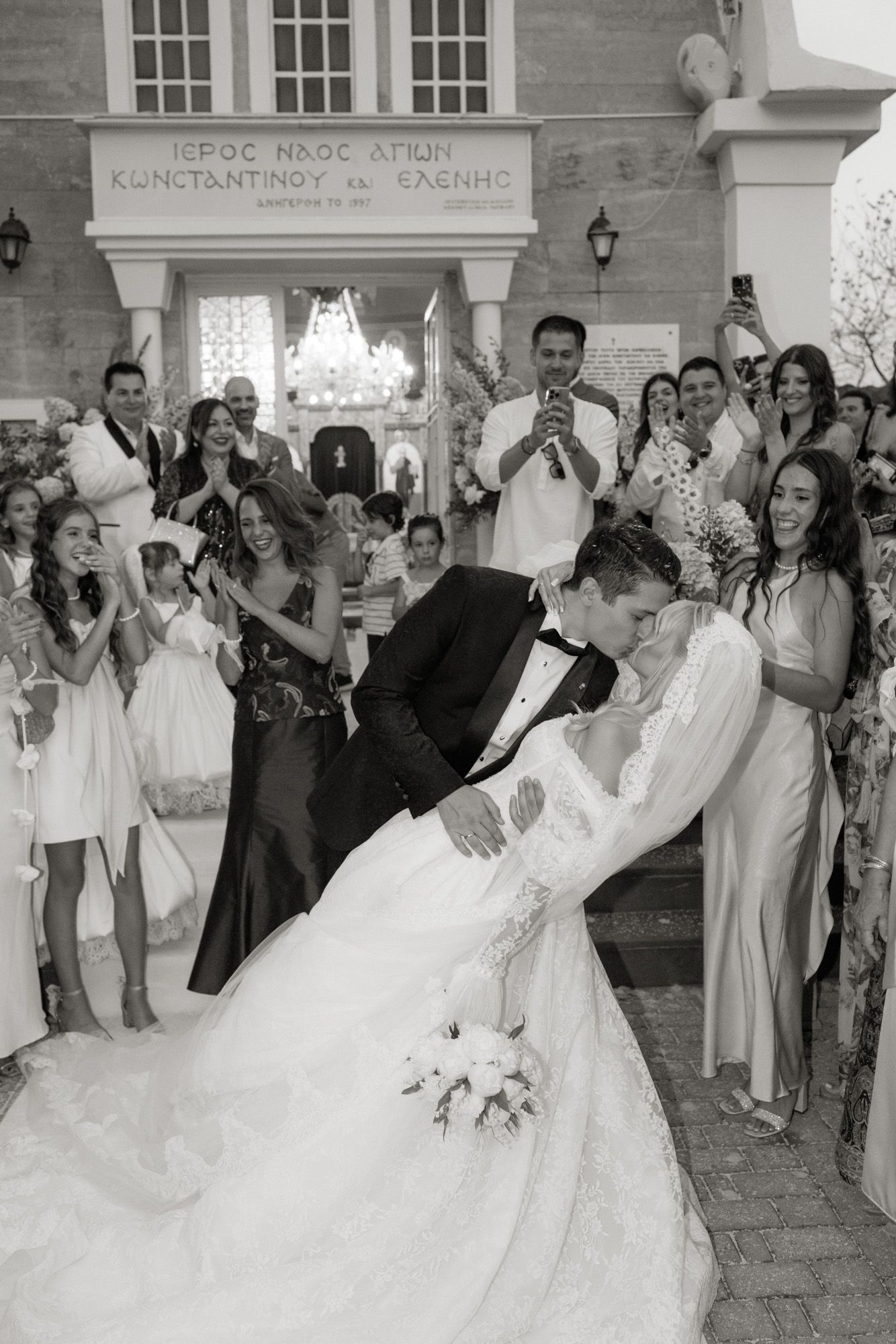 Bride and groom share a romantic dip kiss outside Greek Orthodox church surrounded by cheering wedding guests.