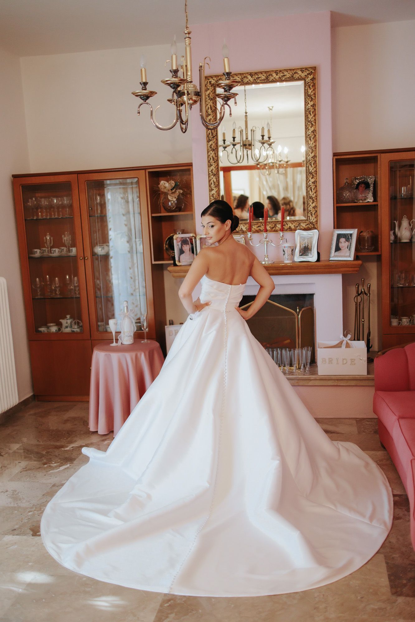 Bride in elegant strapless white ballgown with long train poses in front of ornate gold mirror in decorated room.
